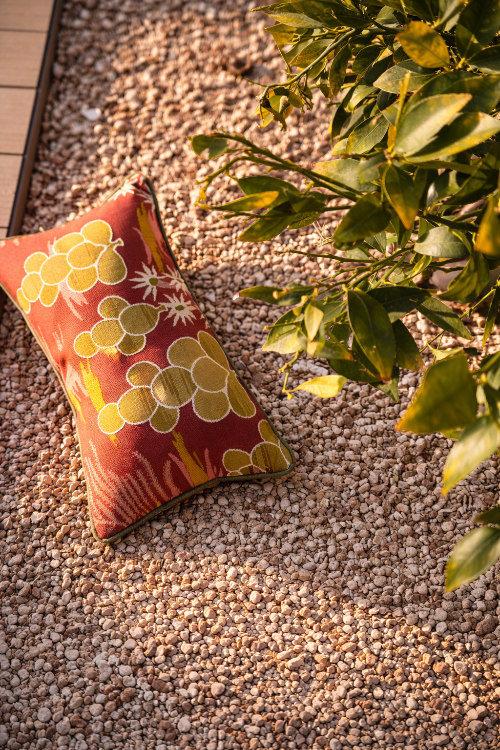 A patterned pillow with grapes design lying on gravel next to a green shrub.