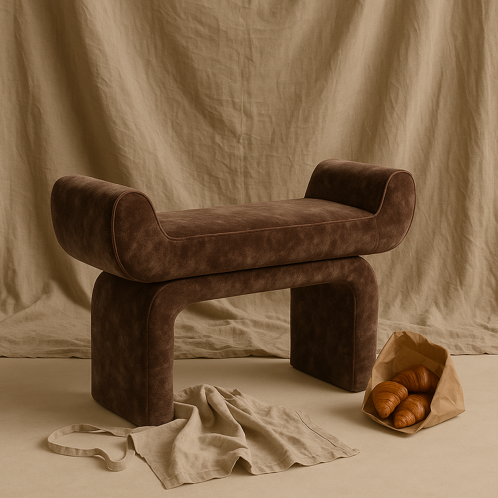 A brown, upholstered bench with curved sides, placed in front of a beige fabric backdrop. A paper bag with croissants is on the floor to the right, with a beige cloth and a strap in front of the bench.