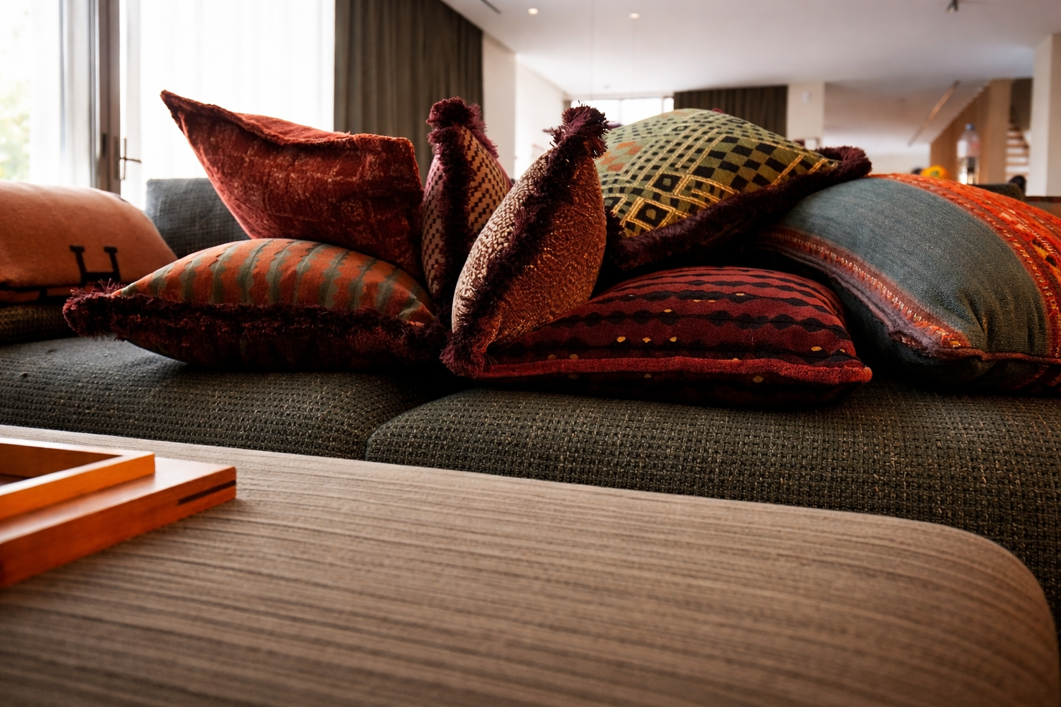 A sofa covered with multiple decorative pillows in various colors and patterns, set in a well-lit room with curtains.