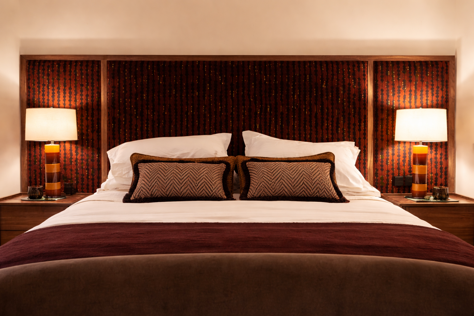 A neatly made bed with white sheets, dark brown blanket, decorative pillows, and a highlighted headboard with a patterned design. There are two lamps on dark wood nightstands on both sides of the bed, with small decor items on each.