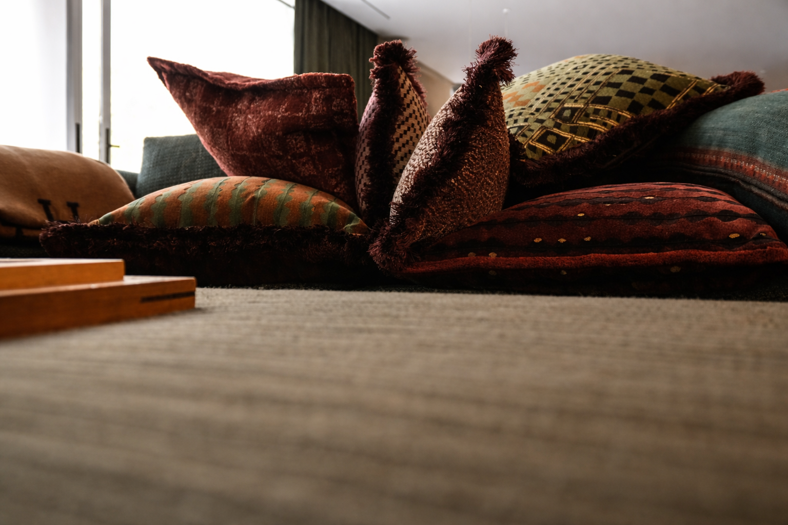 A close-up view of a pile of decorative pillows on a carpeted floor in front of a window with curtains.