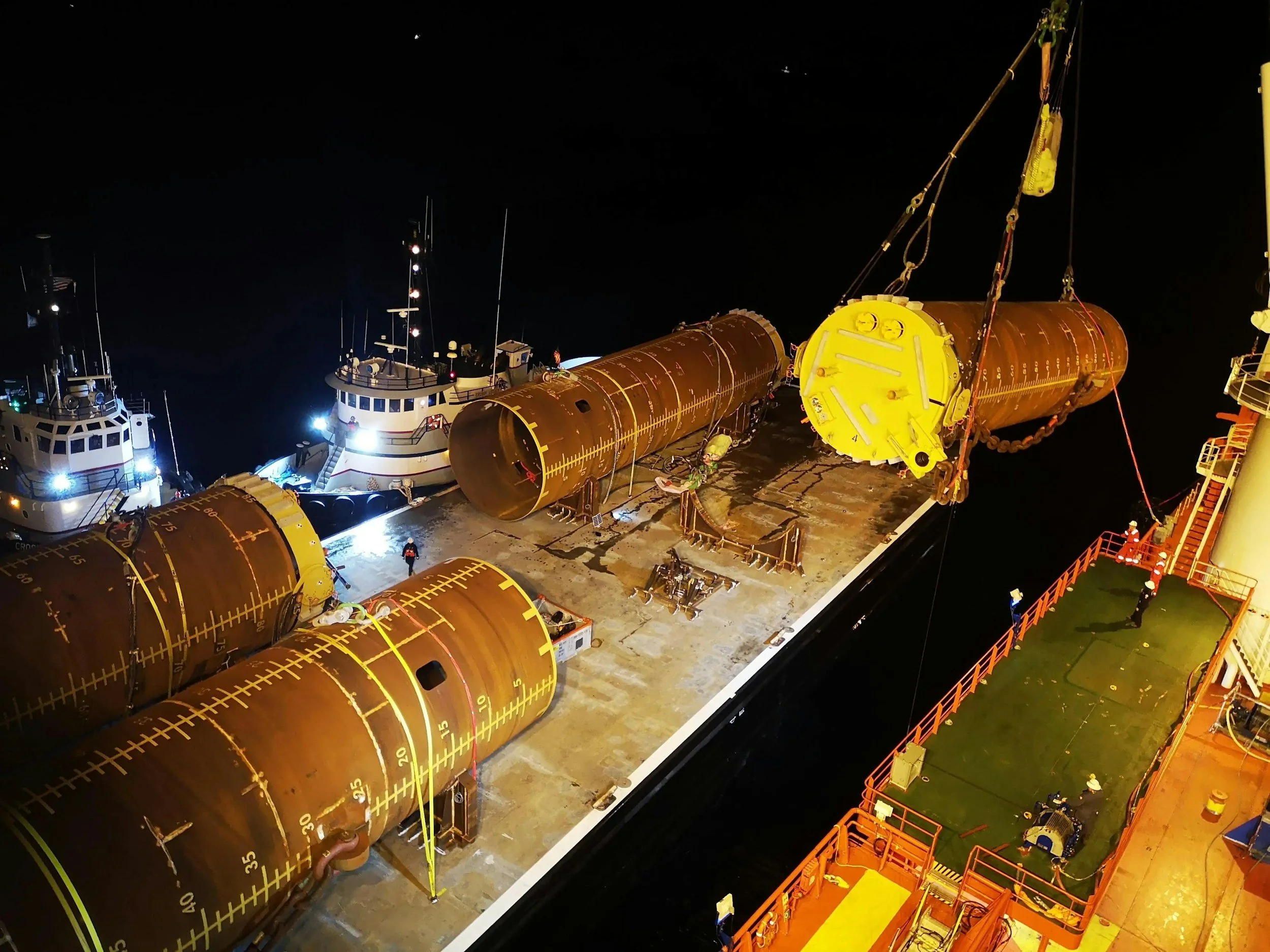 Workers on a ship installing large, cylindrical, rust-colored modules at night, with tugboats nearby.