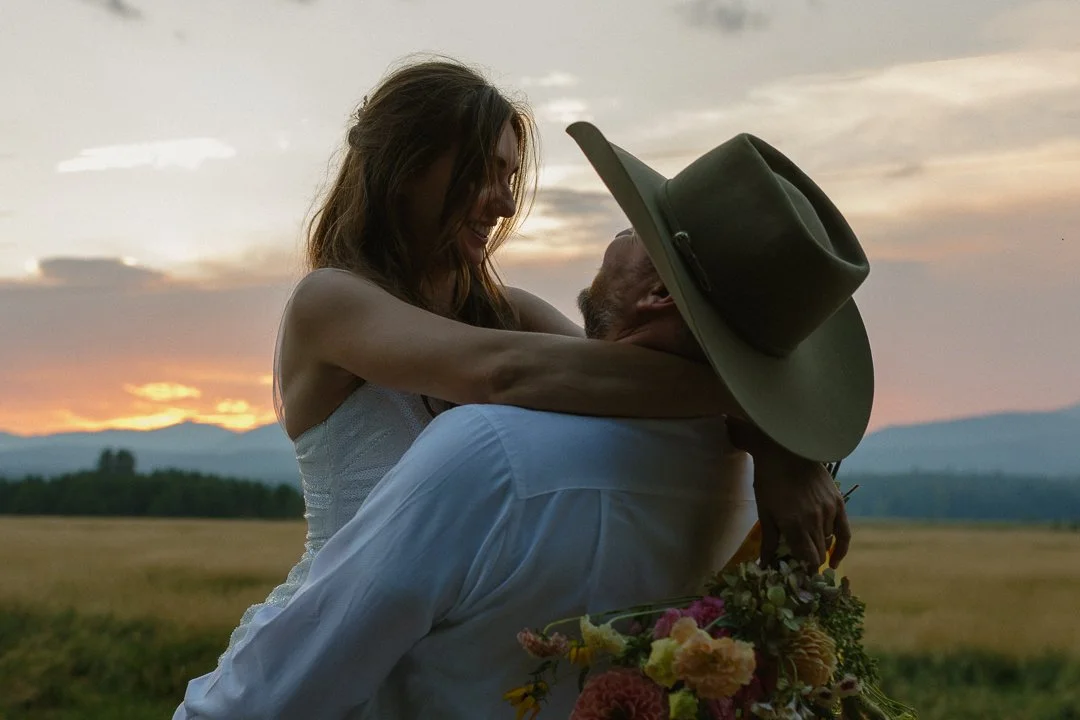 A woman hugging a man outdoors at sunset, with the woman smiling and the man wearing a hat, holding a bouquet of flowers.