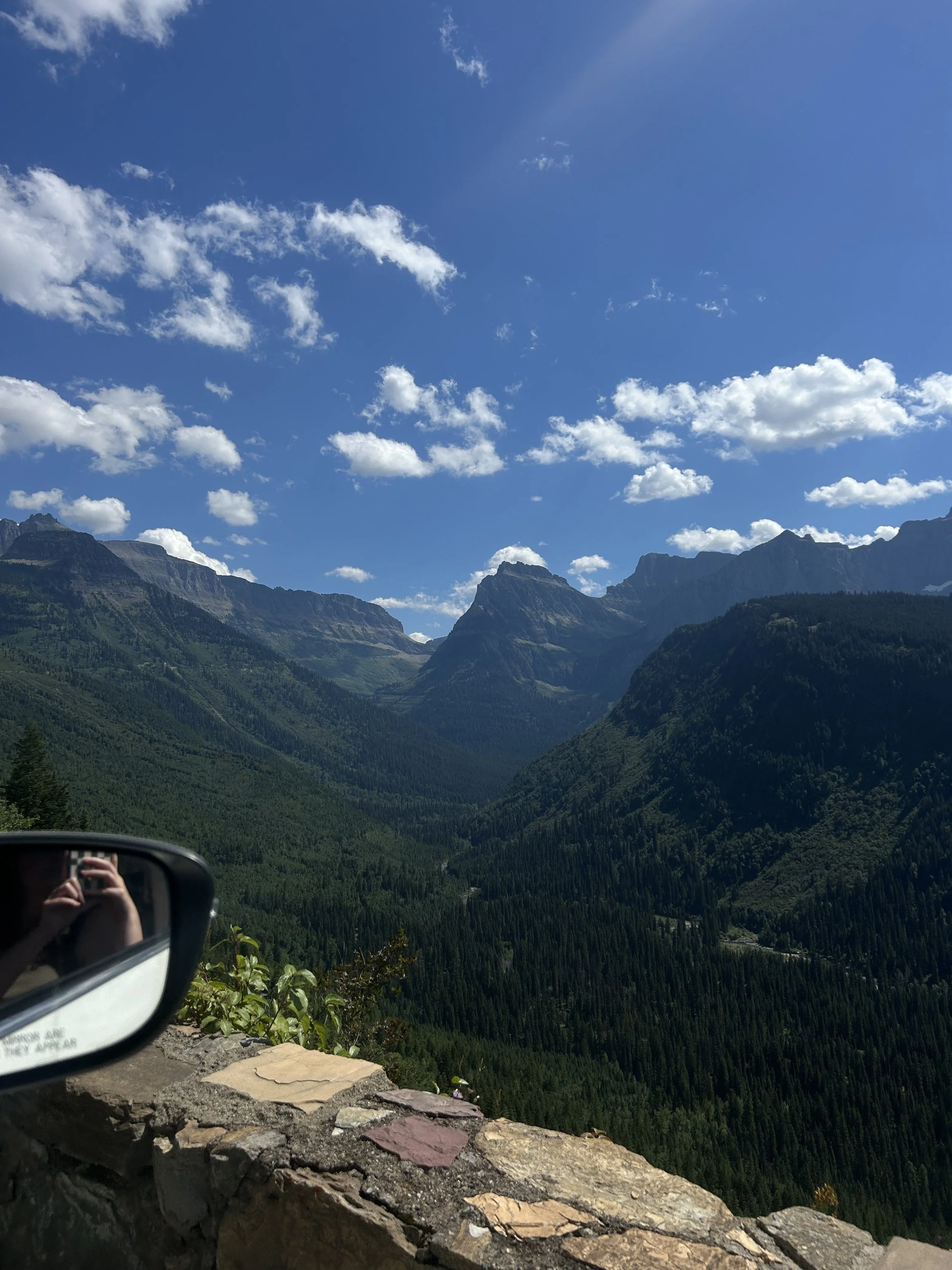 Mountain landscape with blue sky, white clouds, and lush green forested mountain valleys, seen from a car window with a side mirror reflecting the photographer.