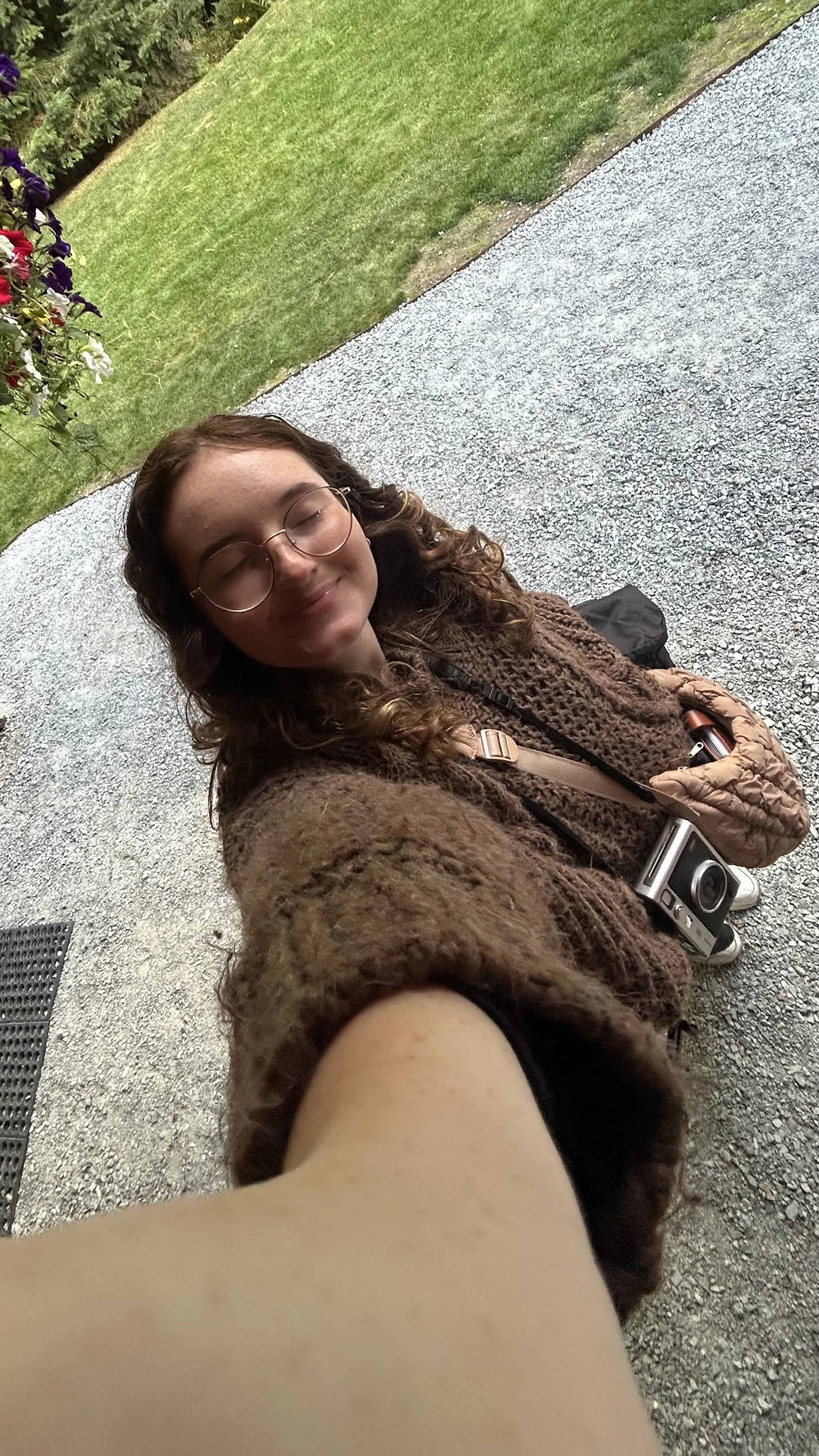 A young woman with glasses and curly hair takes a selfie outdoors, smiling with her eyes closed, wearing a brown sweater. She has a camera hanging around her neck, a small backpack, and a quilted purse on the ground near her. The background includes a gravel pathway, green grass, and some bushes or trees.