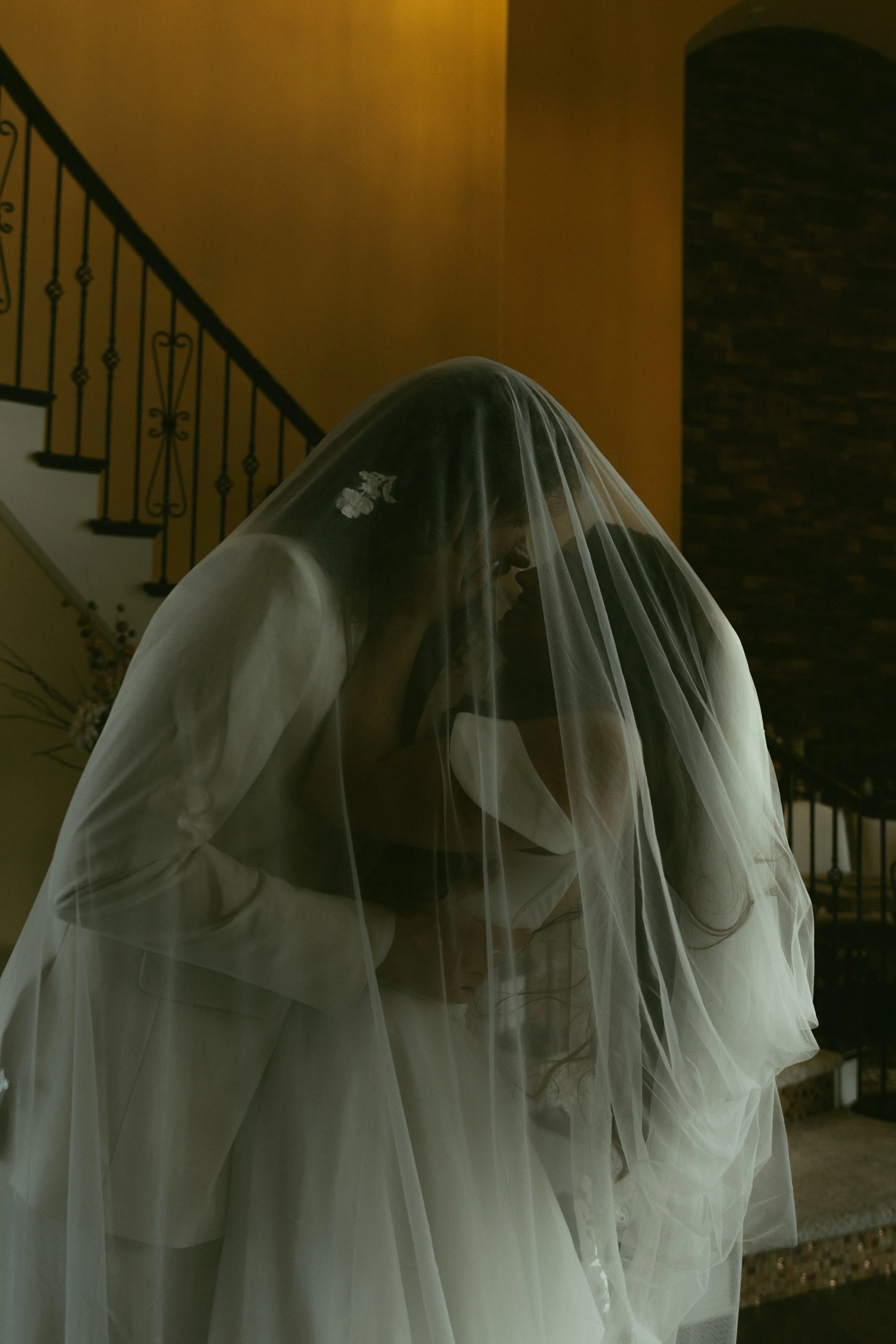 A bride in a white wedding gown with a veil, leaning in to kiss a groom, inside a house with a staircase in the background.