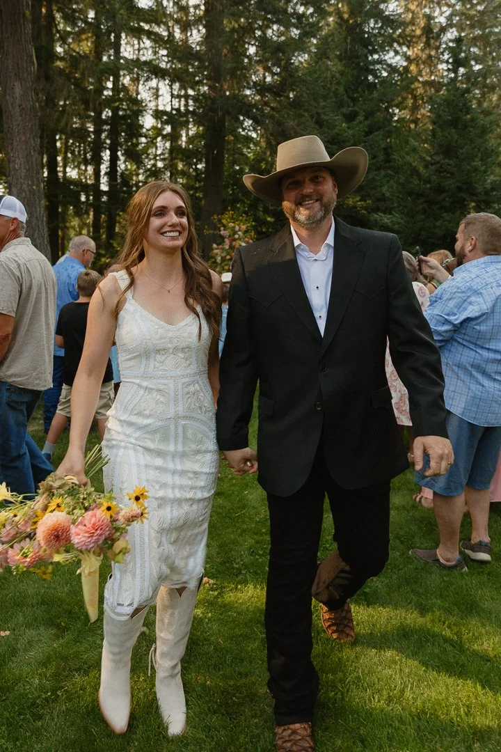 A smiling young woman in a white dress holds a bouquet of flowers while walking hand-in-hand with a smiling man in a black suit and large cowboy hat at an outdoor event surrounded by other people and trees.