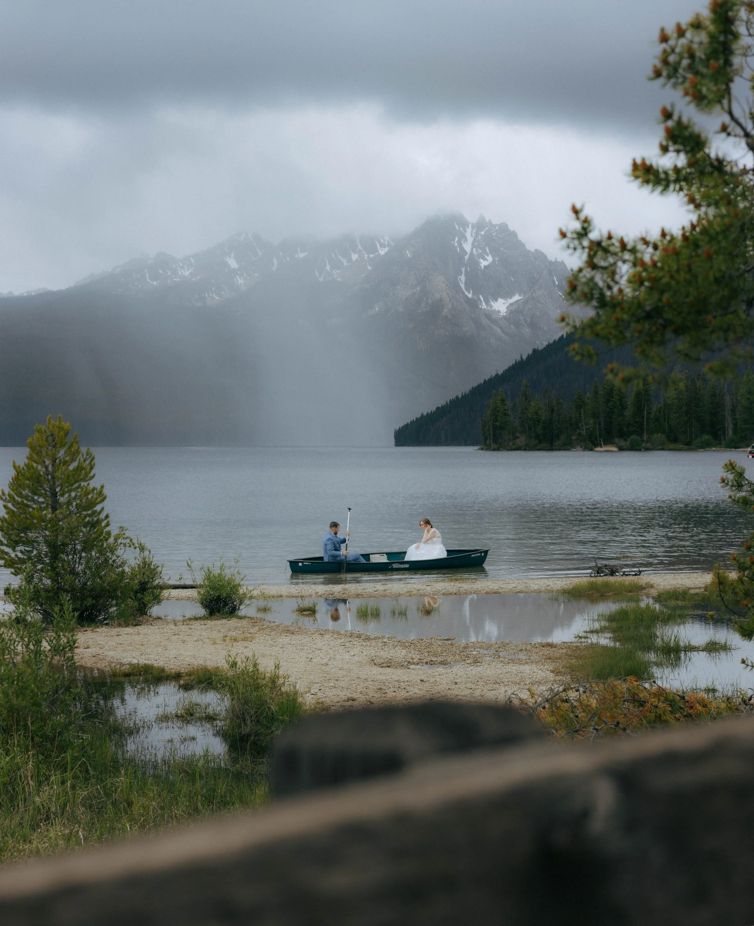 A couple in a boat on a lake, with snow-capped mountains and a cloudy sky in the background, and some trees and bushes along the shoreline.