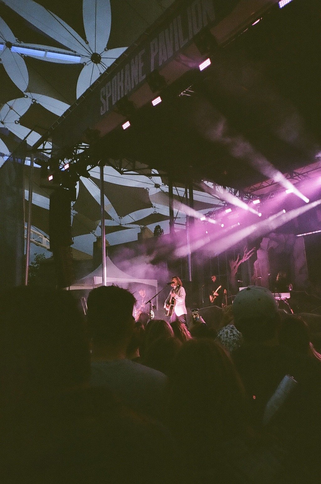 A live outdoor concert at sunset featuring a man playing guitar on stage, with purple stage lights and an audience in the foreground. Stage is under a canopy with a sign reading 'Spokane Pavillion'