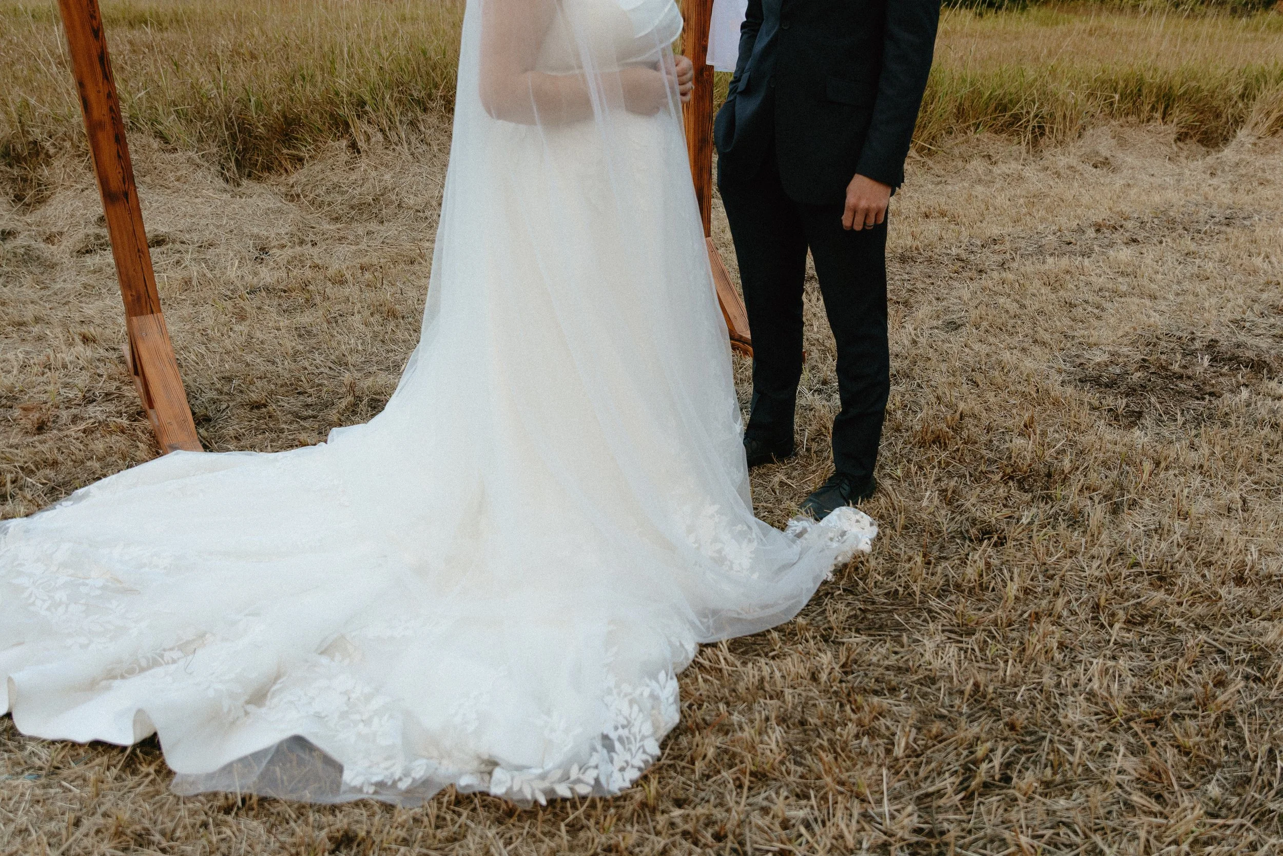 A bride in a white wedding dress and a groom in a black suit standing outdoors on dry grass near a wooden wedding arch.