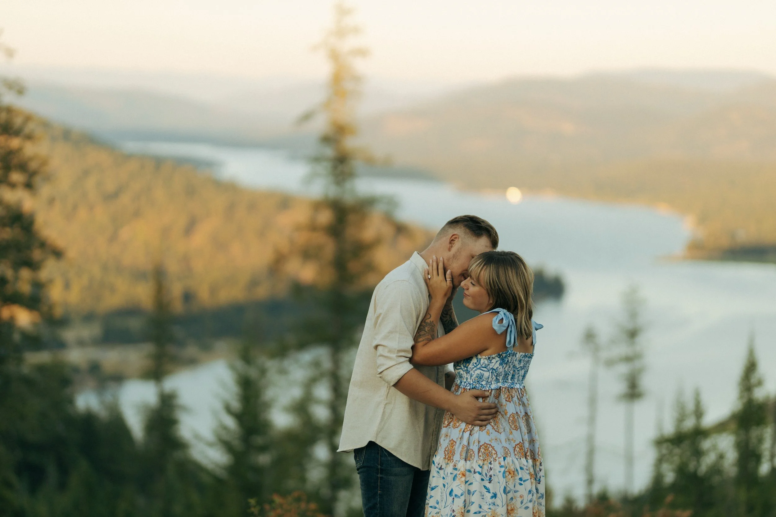 A couple standing close together outdoors by a body of water, sharing an intimate moment during sunset or late afternoon, with trees and a distant landscape in the background.