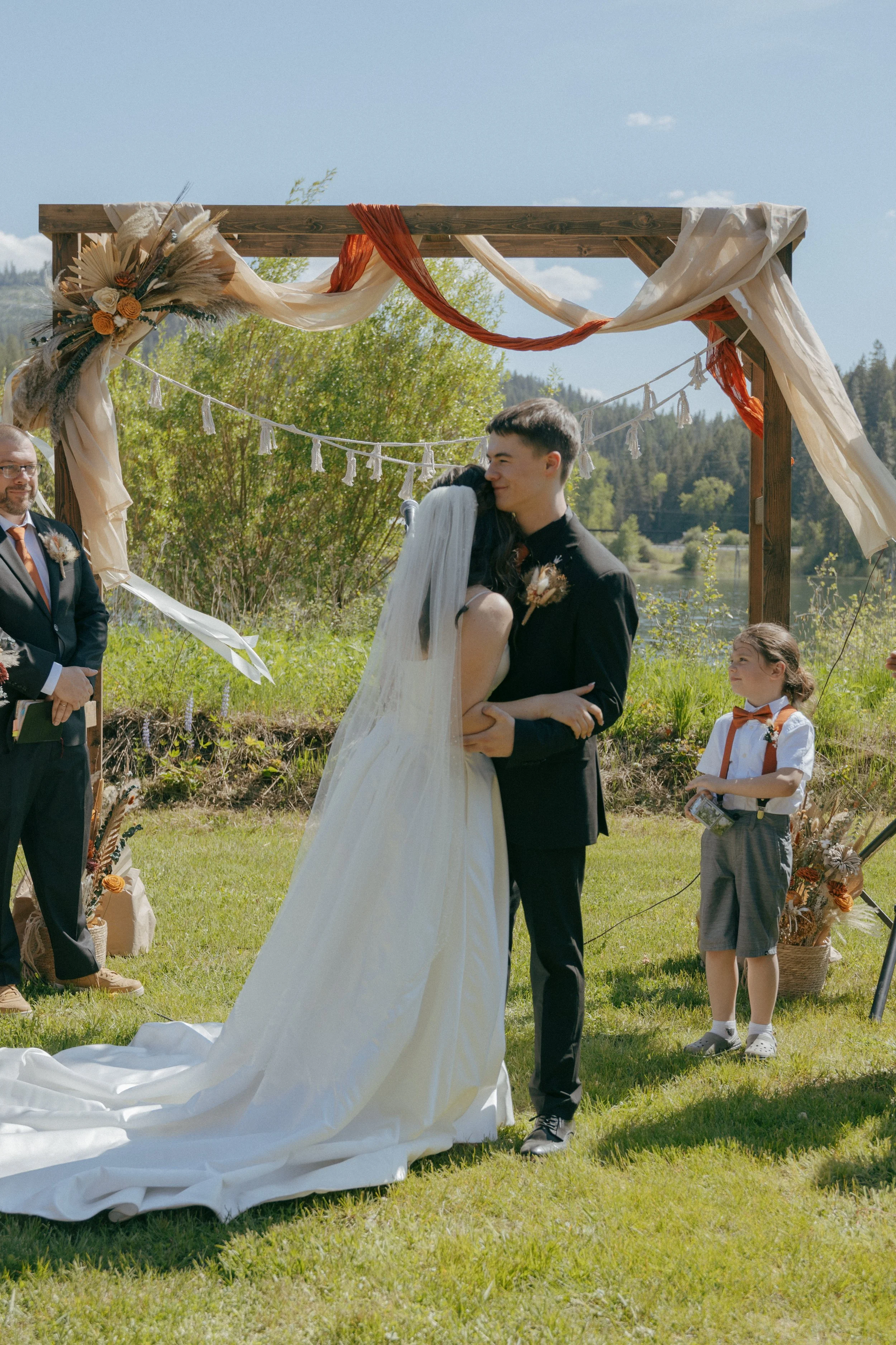 A couple getting married outdoors under a decorated wooden arch, with a man in a suit, a young girl, and a scenic river and trees in the background.