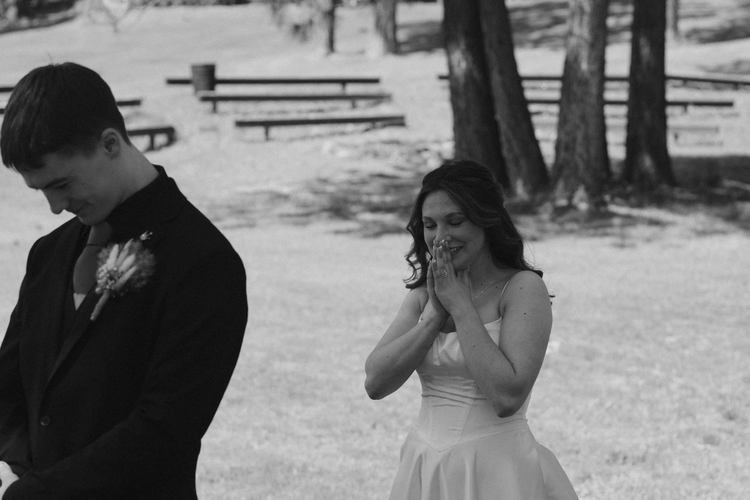 A woman in a wedding dress praying or holding her hands together with a smile, standing outdoors near trees, while a man in a suit with a boutonnière stands nearby with his head bowed.