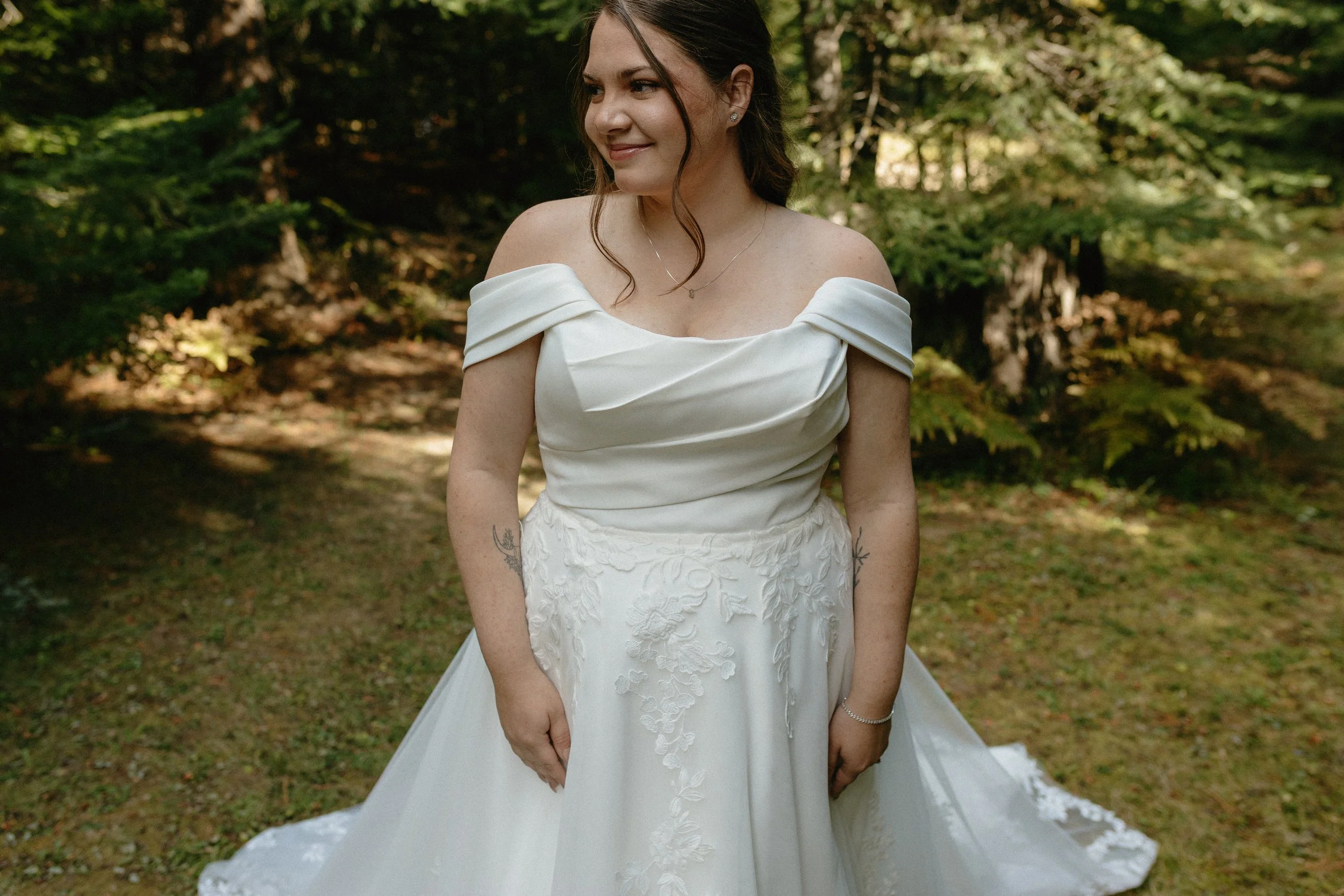 Bride in a white wedding dress standing outdoors in a wooded area with green trees background.