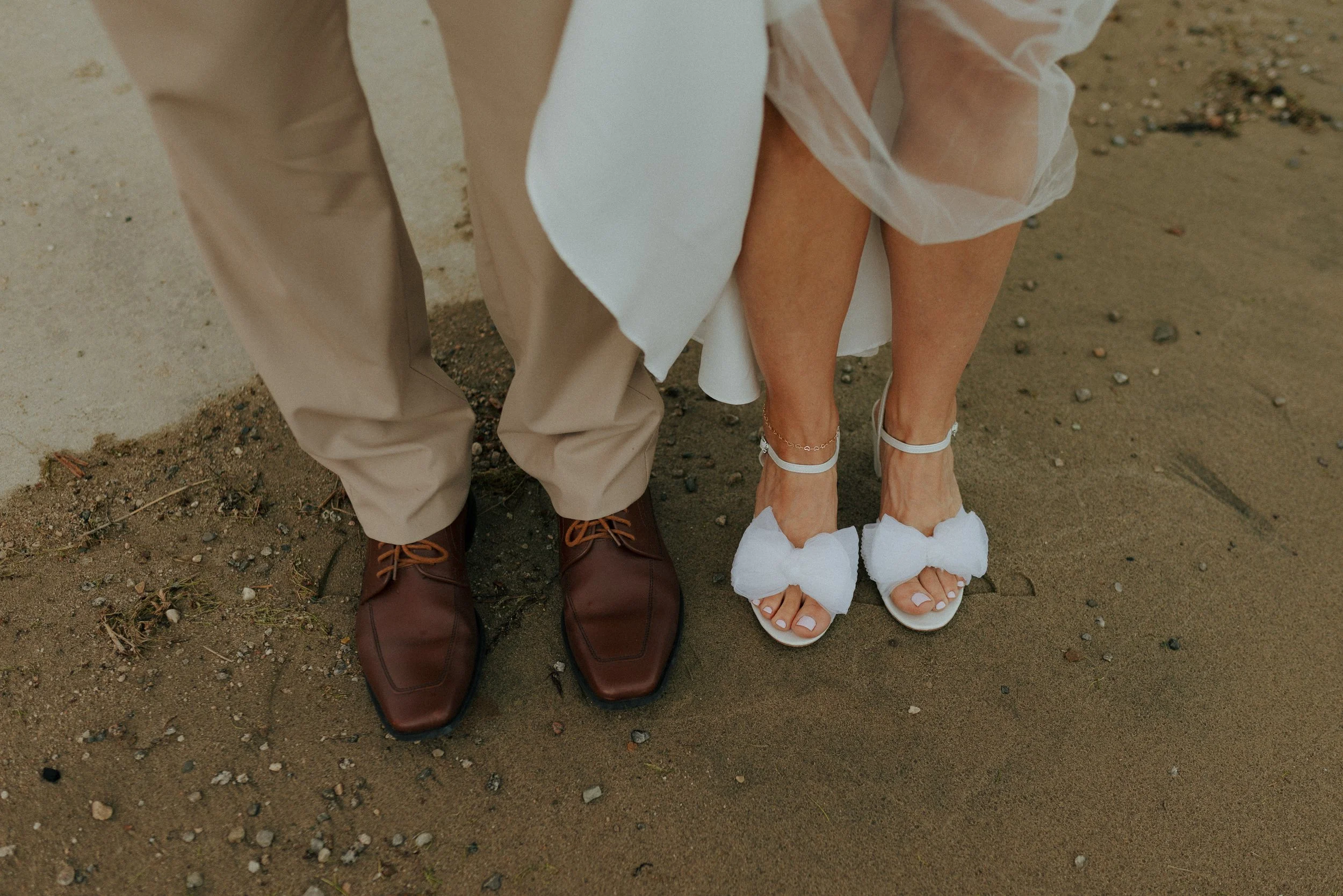 Close-up of a bride and groom's feet on a sandy beach. The groom is wearing beige pants and brown leather shoes. The bride is wearing white wedding sandals with bows and a white dress.