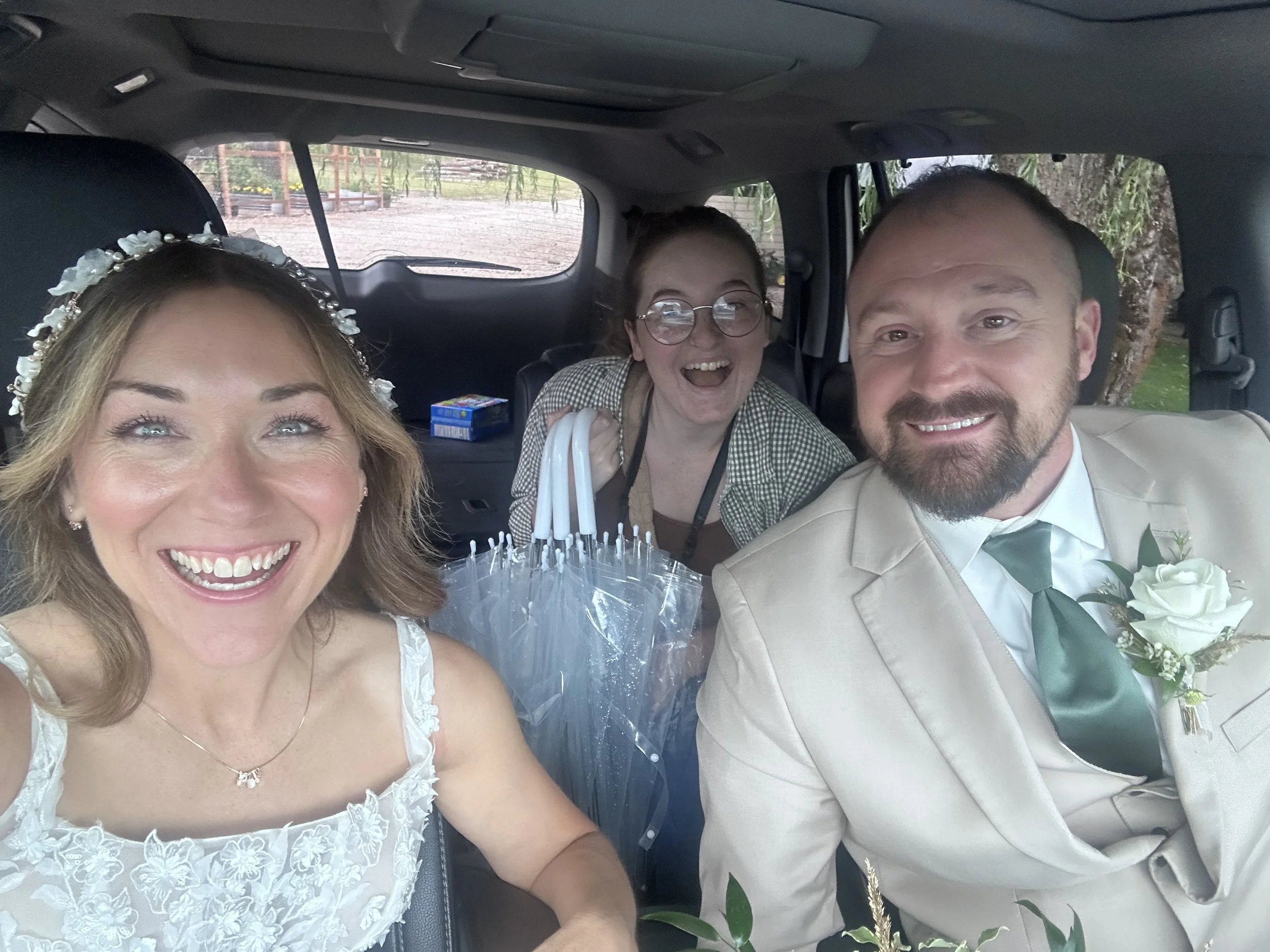 A happy bride, groom, and a woman smiling in the backseat of a car, with the bride wearing a white dress and floral headband, the groom in a beige suit with a boutonnière, and the woman in glasses holding umbrellas.