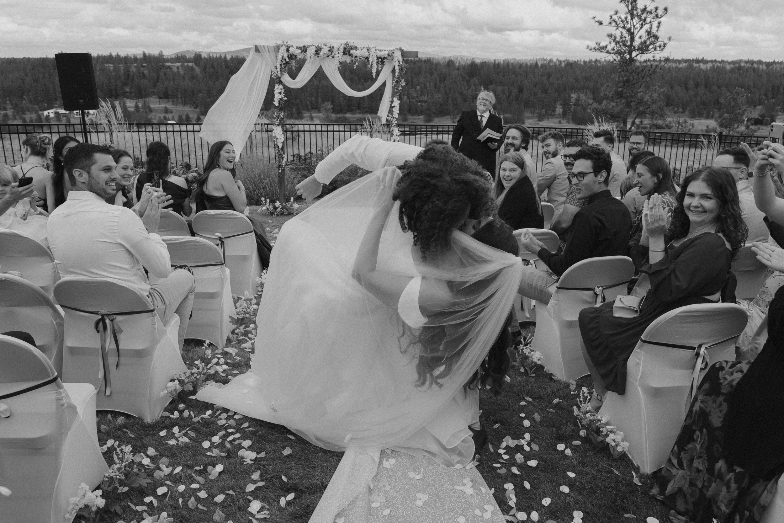 Black and white photograph of a wedding ceremony outdoors with a bride and groom kissing under a floral archway, surrounded by seated guests clapping and smiling, on a grassy area with flowing chairs and flower petals on the ground.