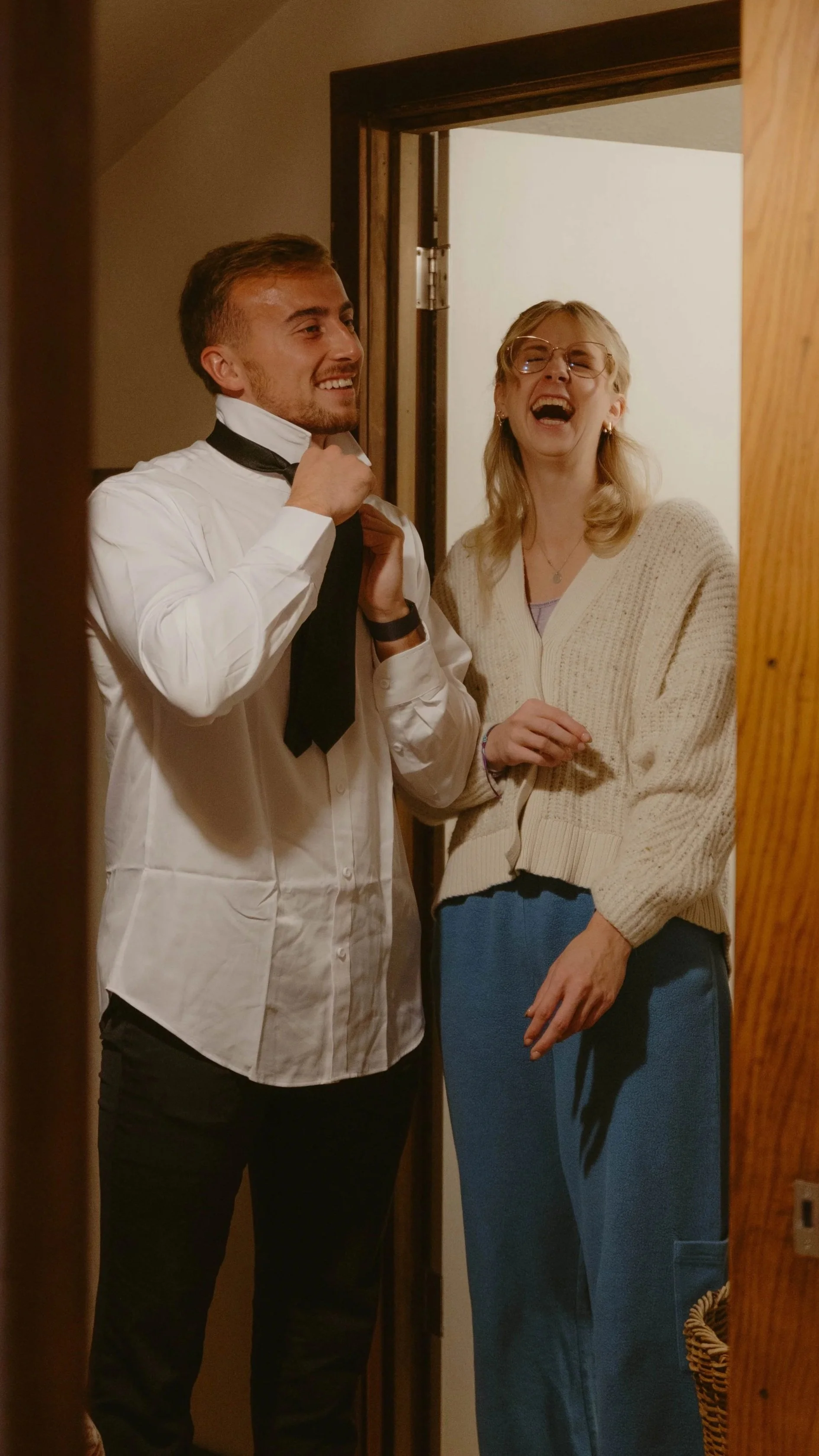 A young man fixing a bow tie on a woman, both smiling and laughing, standing in a doorway.