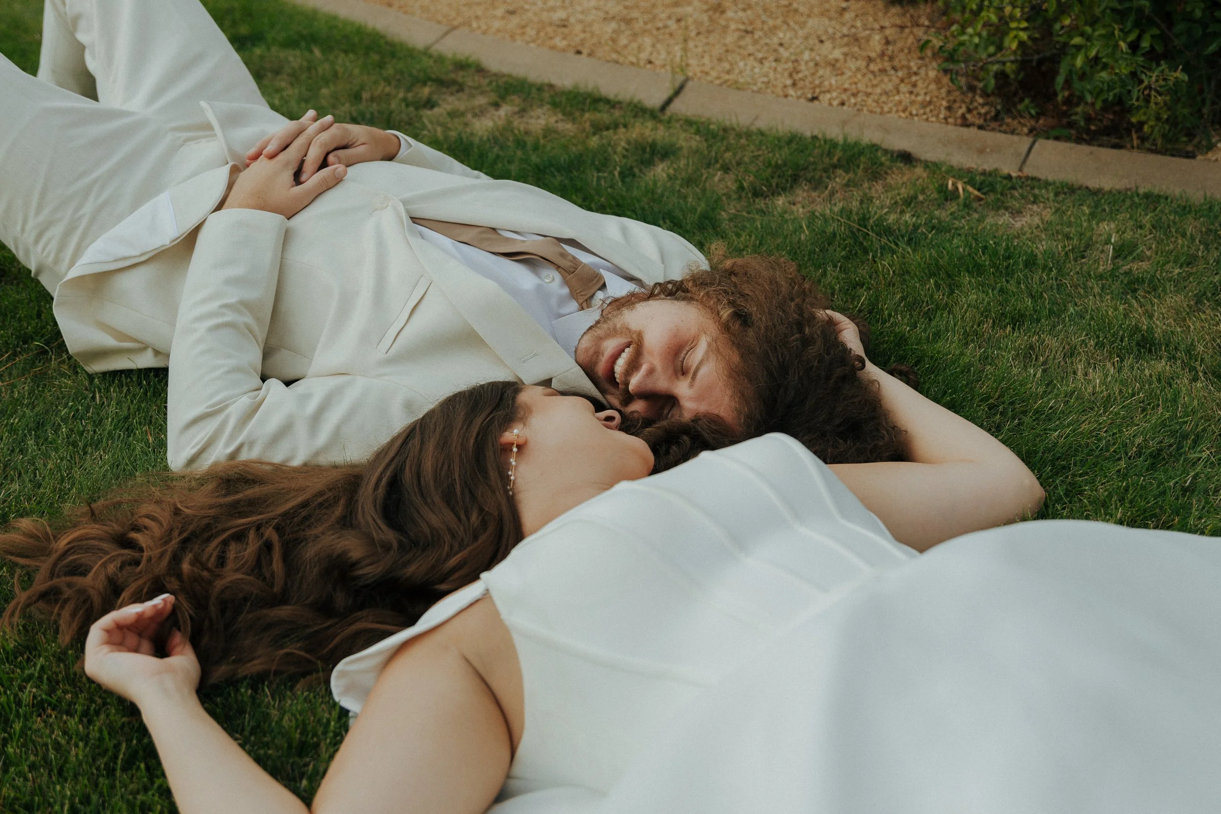 A couple lying on the grass outdoors, smiling and looking at each other, with the woman holding the man's head.