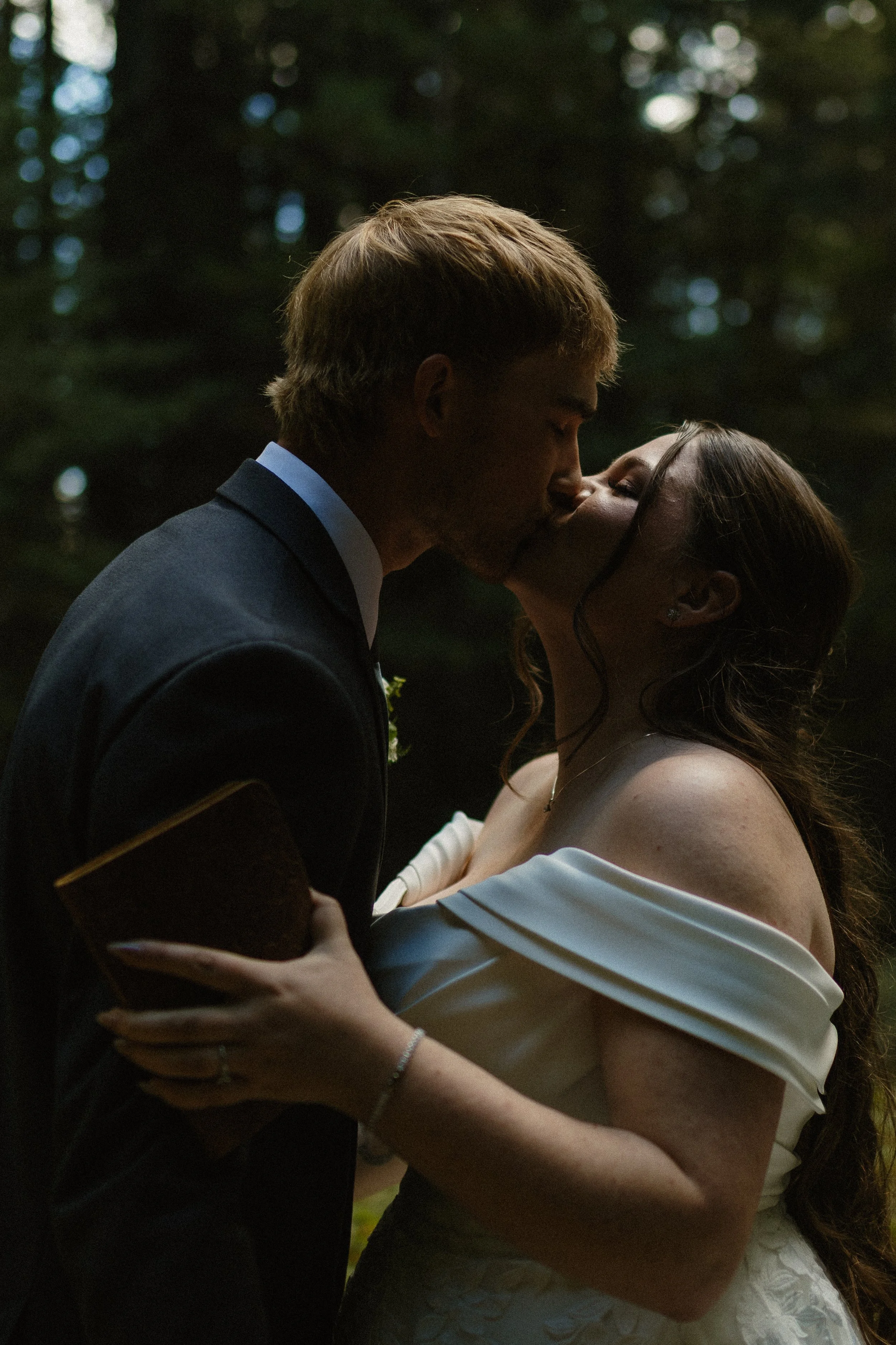 A couple sharing a kiss outdoors, likely during a wedding or special occasion, with a dark forest background.
