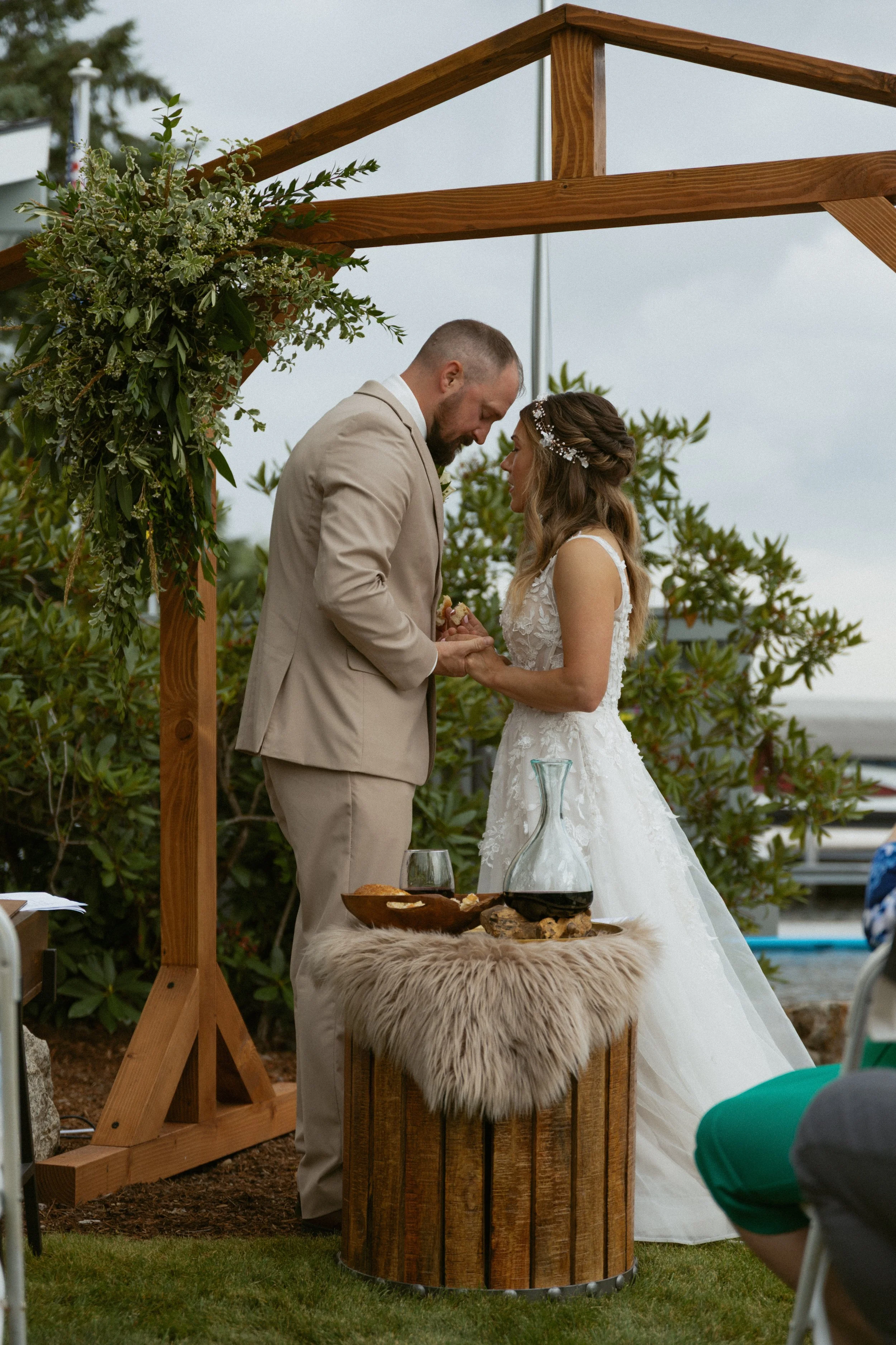 Bride and groom exchanging rings during outdoor wedding ceremony under a wooden arbor with floral decorations.