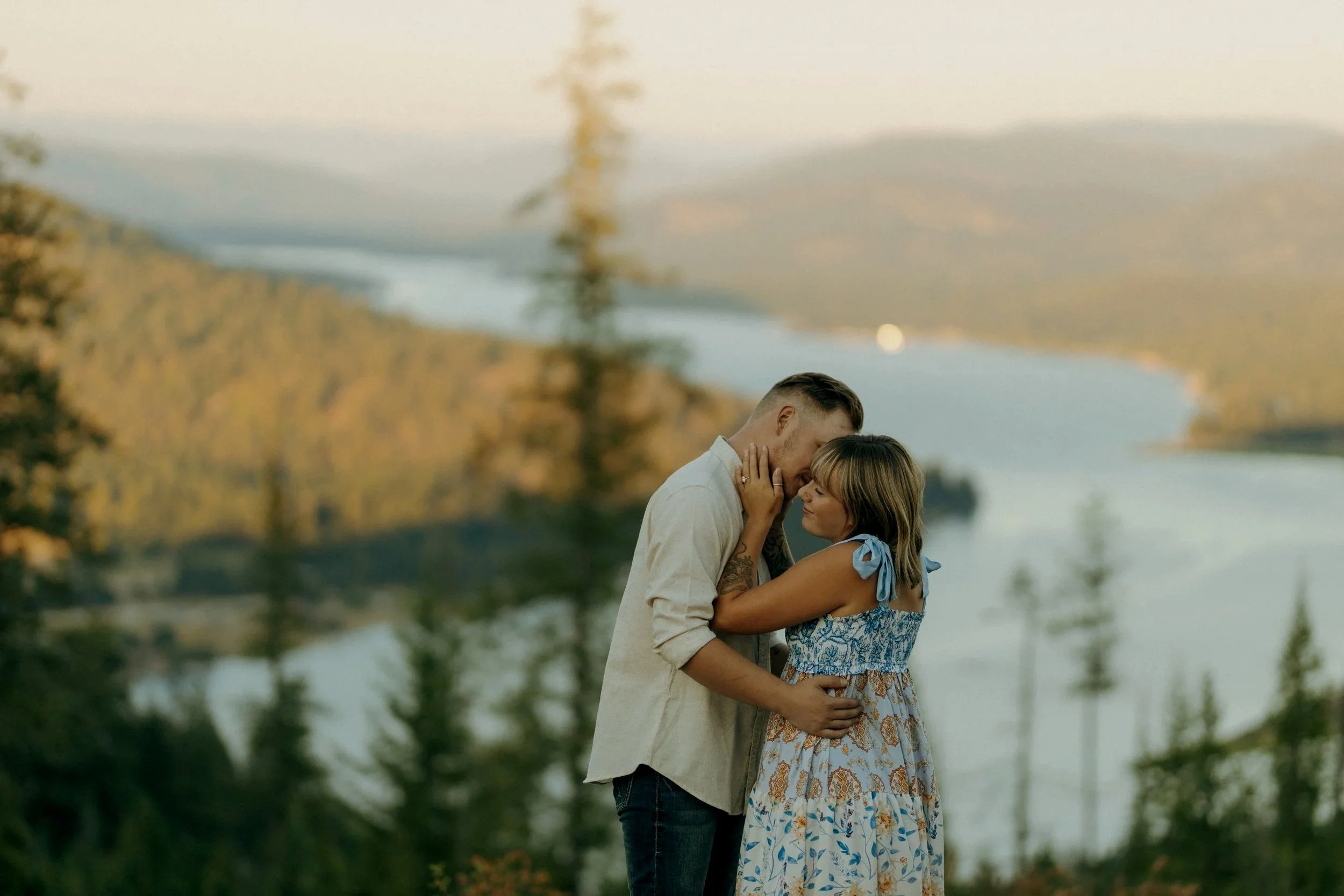 A man and woman embrace near a body of water with trees and hills in the background during sunset.