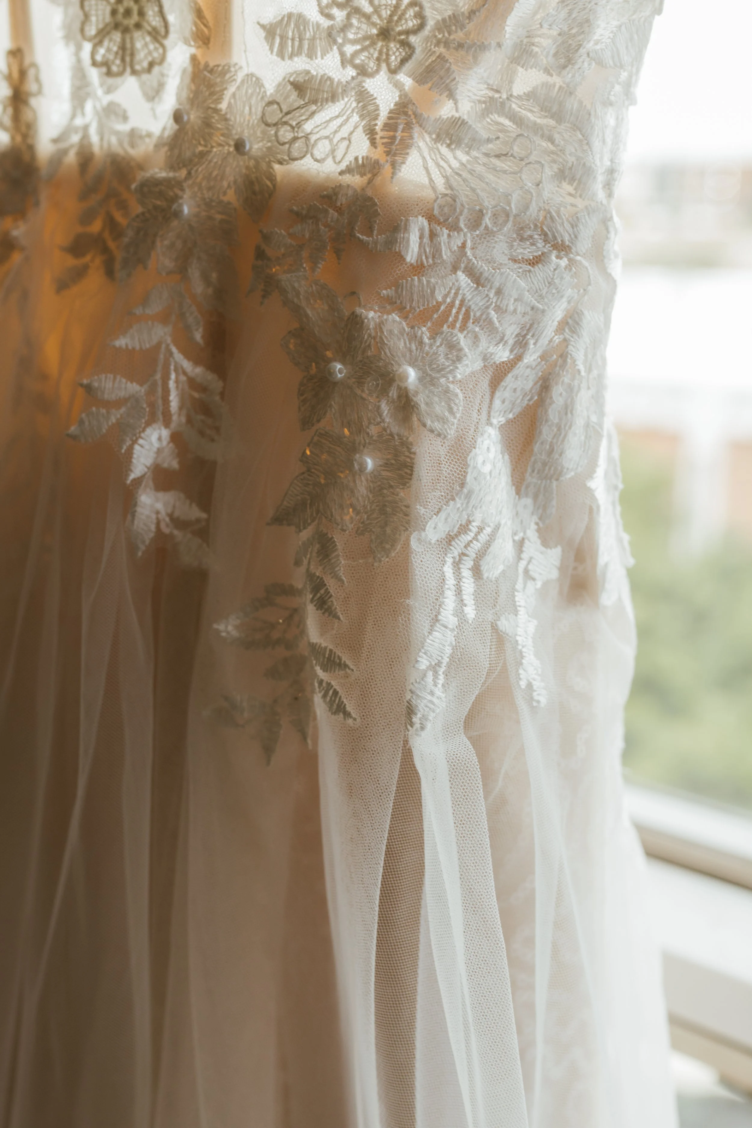 Close-up of a delicate lace curtain with floral embroidery and pearl accents, hanging in front of a window with natural light.
