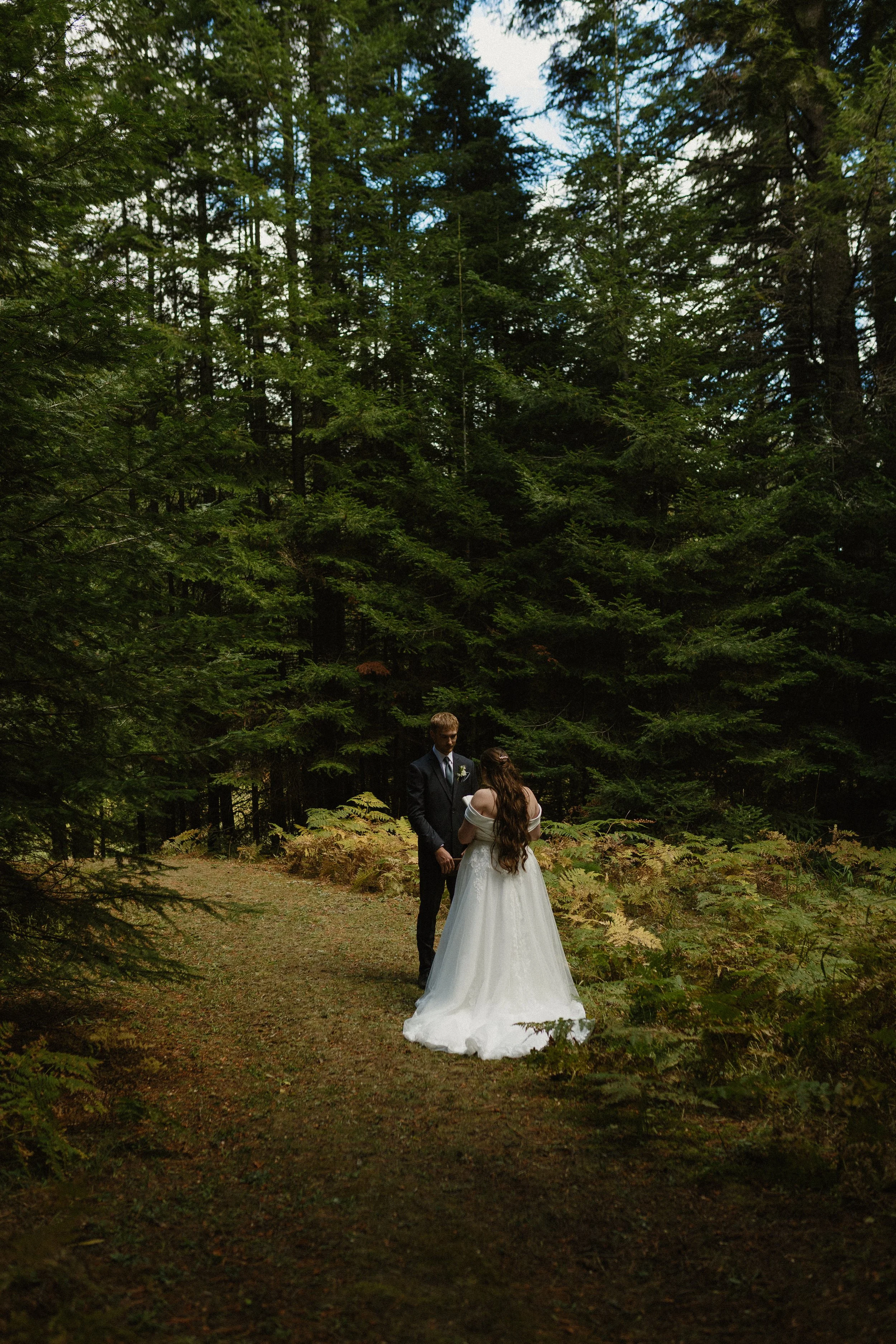 A bride and groom standing hand in hand on a forest pathway during their wedding, surrounded by tall green trees and foliage.