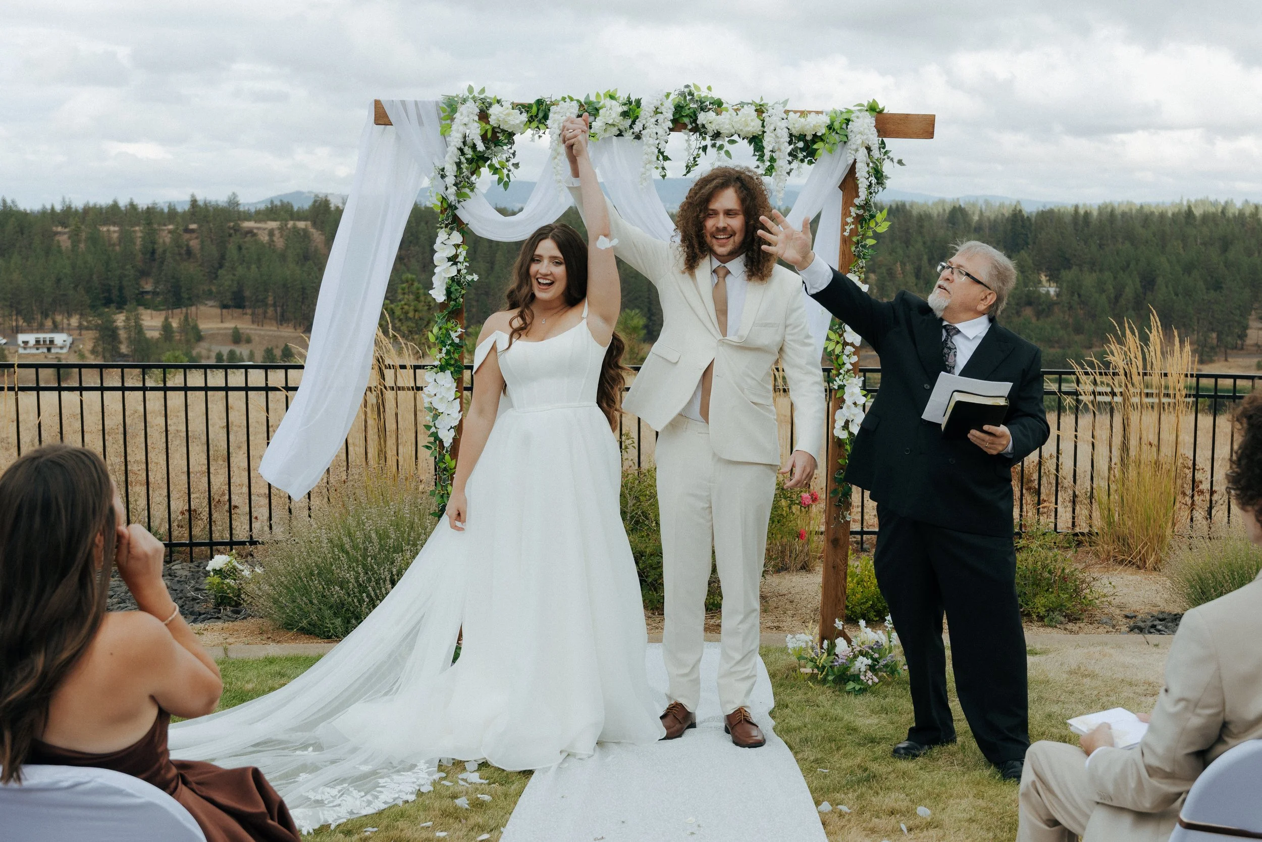 A bride and groom celebrate their wedding under a decorated outdoor arch with a officiant, as guests watch.