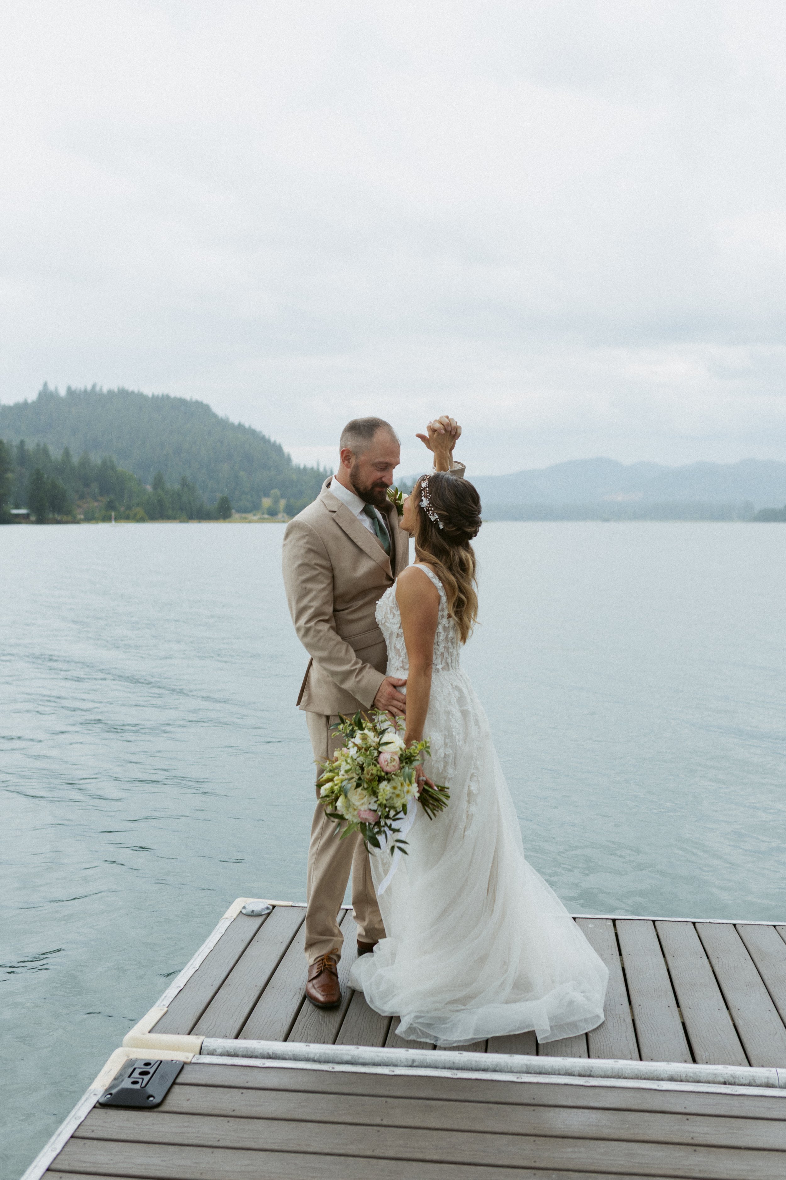 A bride and groom embrace on a dock by a lake, with mountains in the background. The bride holds a bouquet and wears a white wedding dress, while the groom is dressed in a tan suit.