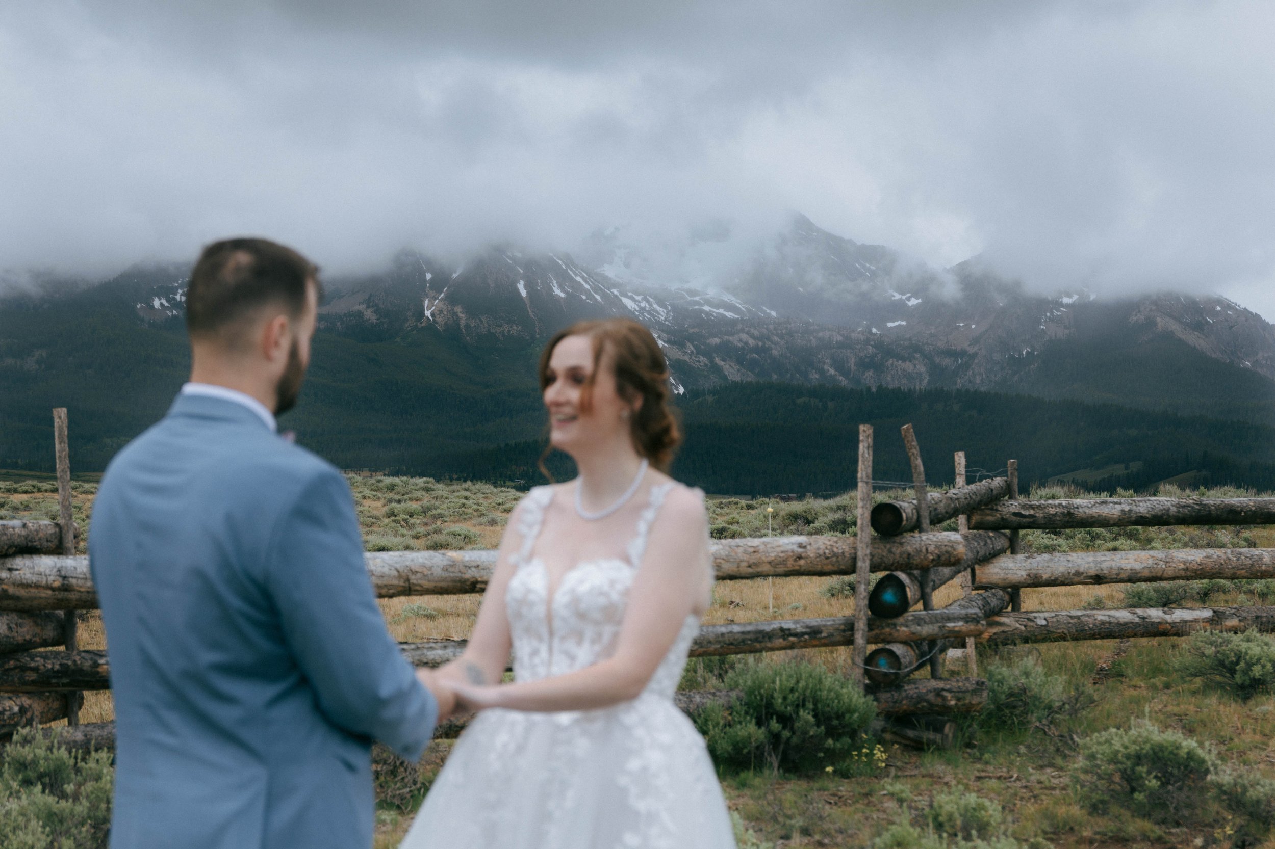 A man and woman in wedding attire holding hands outdoors with a mountain range and cloudy sky in the background.