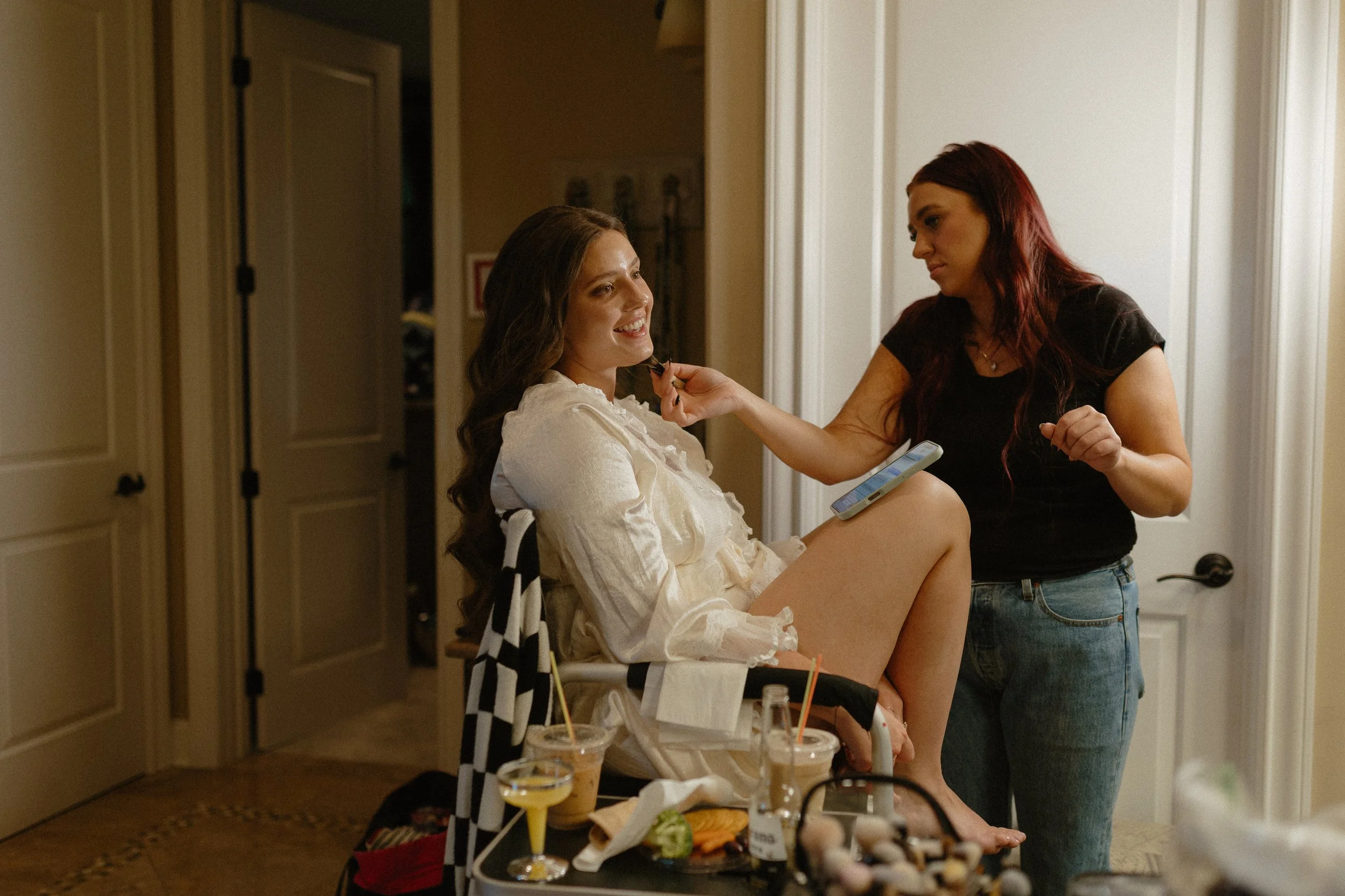 A woman sitting and smiling while another woman applies makeup to her face in a room with makeup and drinks on a table.