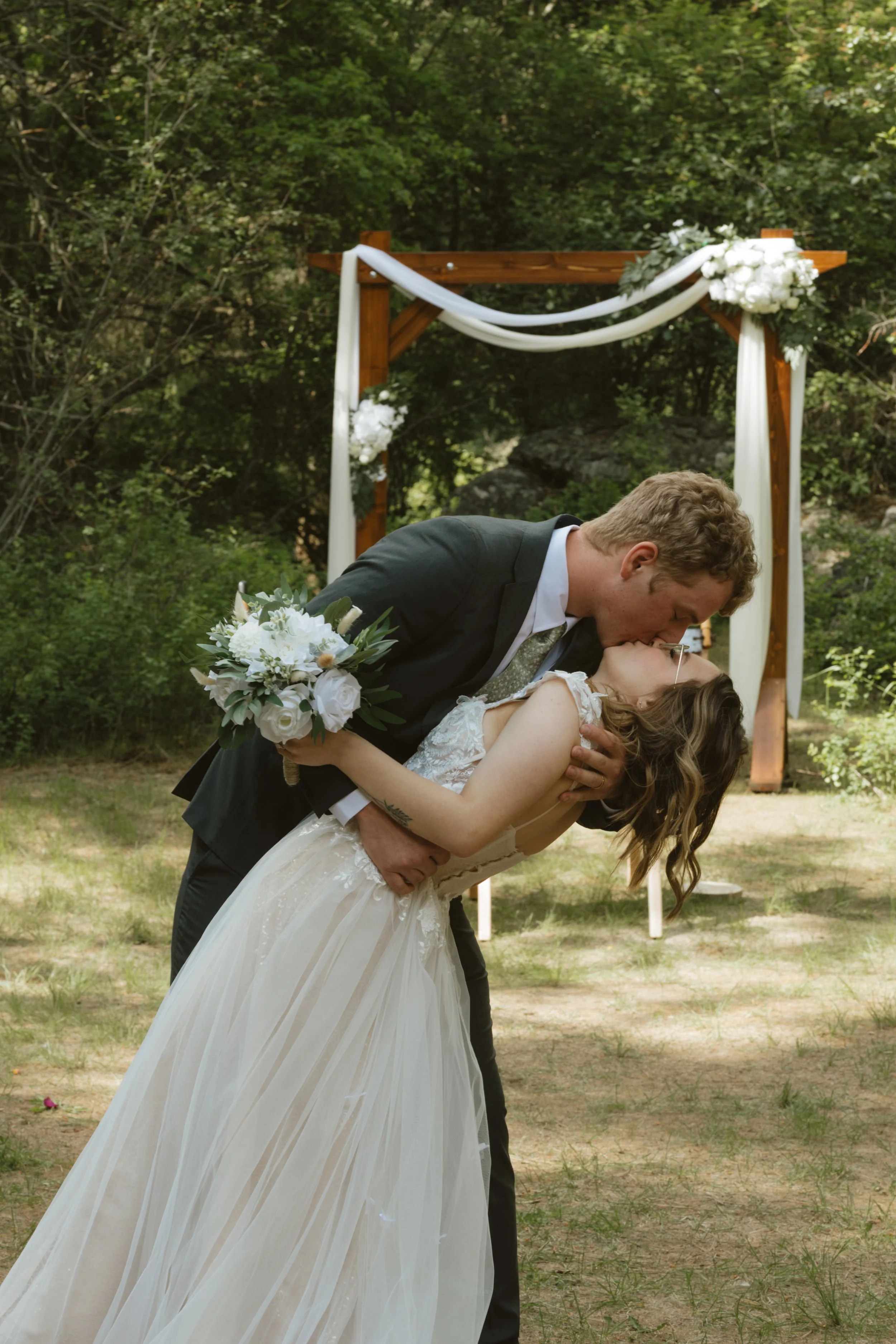 A bride and groom kissing during their wedding ceremony outdoors, with a decorated wooden arch in the background surrounded by greenery.