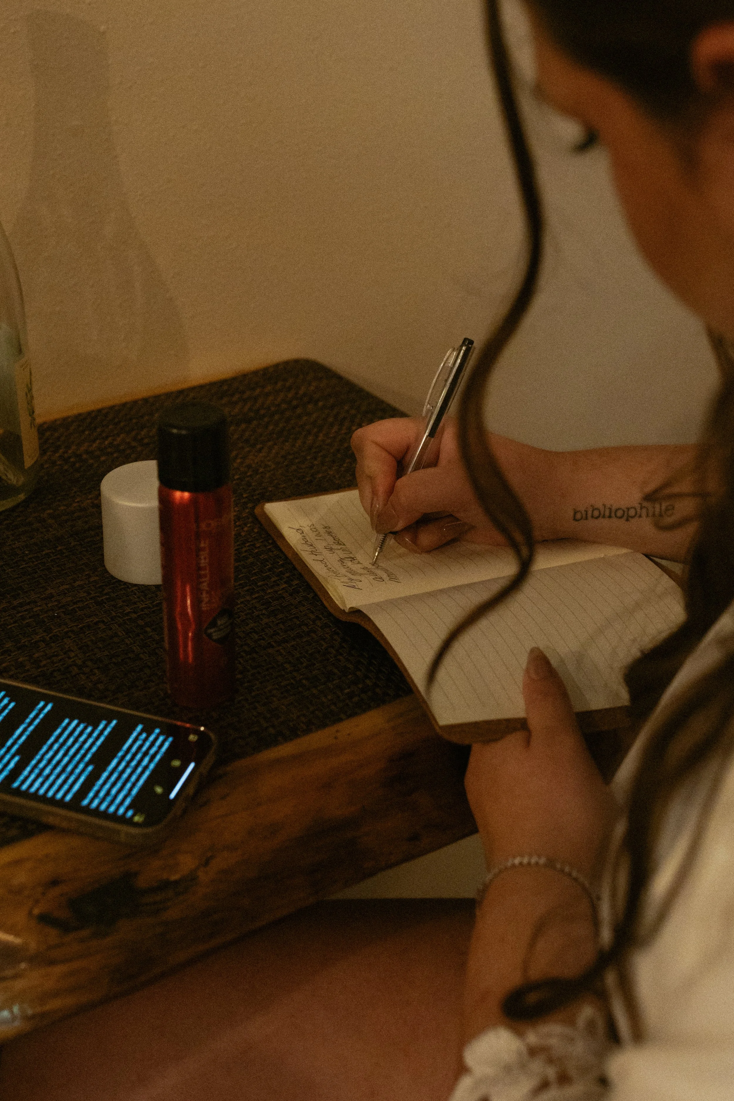 A person writing in a notebook on a wooden table, with a phone displaying blue text, a red bottle, and a white container nearby.