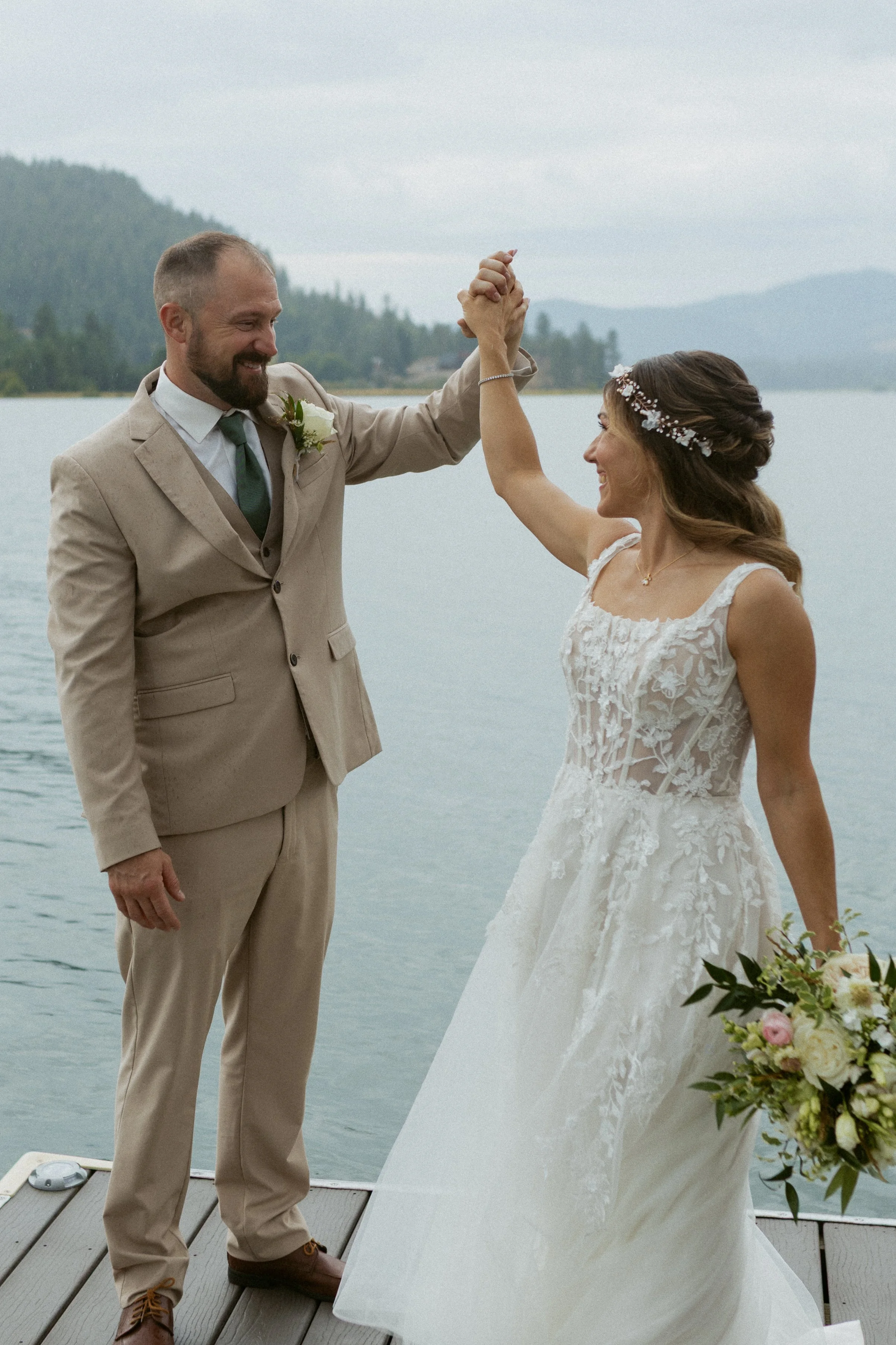 A bride and groom stand on a dock by the water, holding hands up in celebration of their wedding.