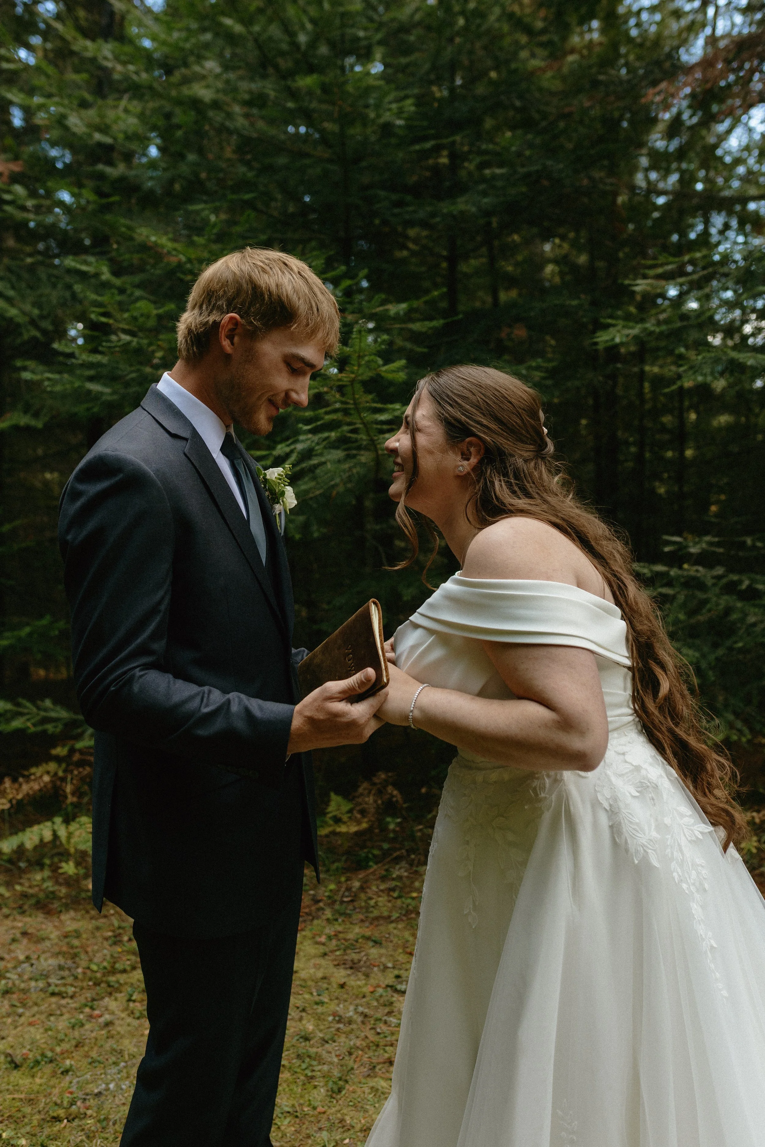 A bride and groom exchanging vows outdoors in a wooded area, with the groom holding a small book or card and both smiling at each other.