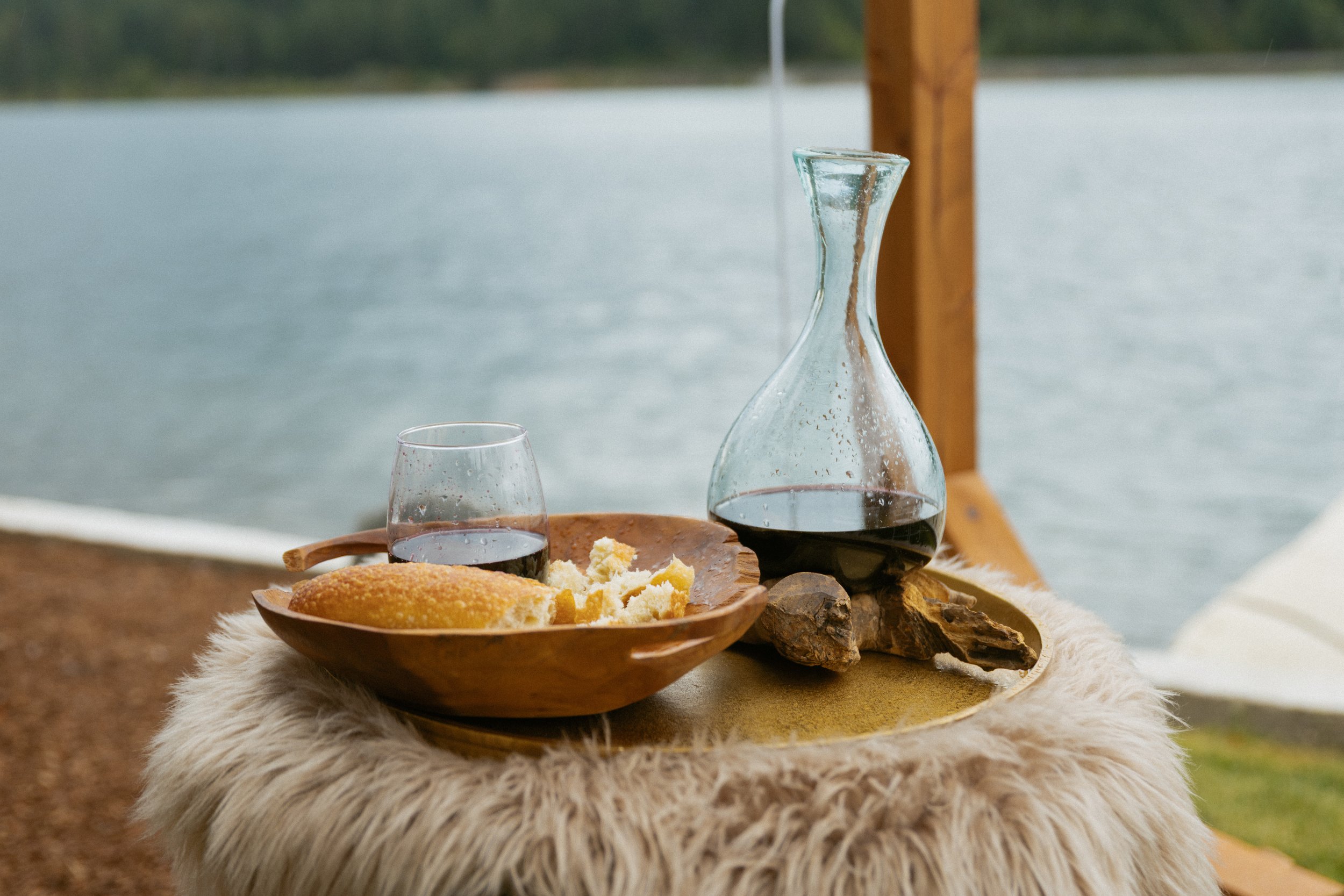 A wooden tray with a glass of red wine, a wine decanter, a plate with bread and bread crumbs, placed on a furry beige mat by a lakeside with water and trees in the background.