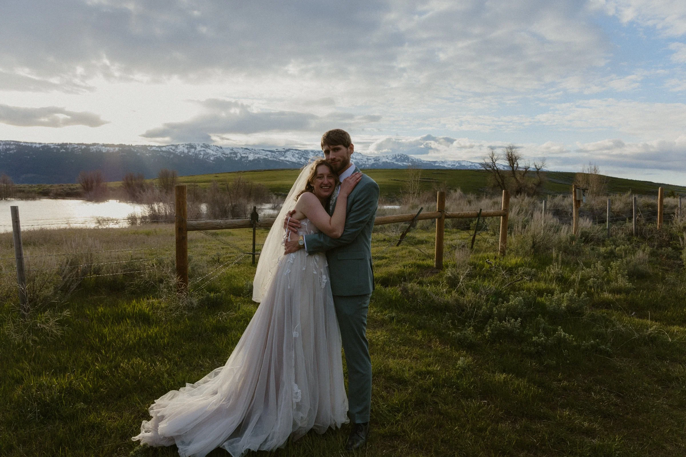 A newlywed couple embracing outdoors in a grassy field with a fence, a lake, and snow-capped mountains in the background, under a cloudy sky.