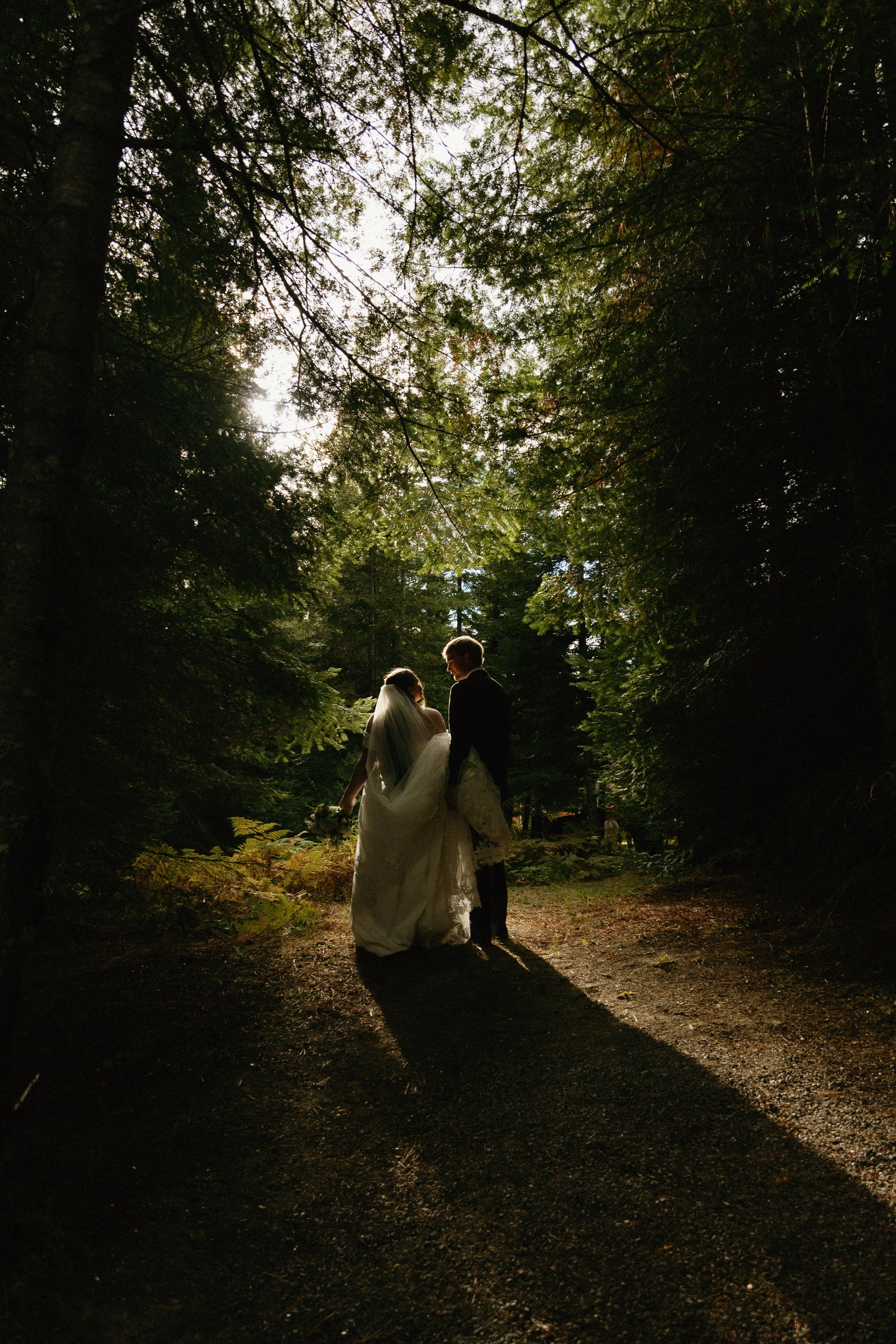 A bride and groom walking through a forested area during sunset.