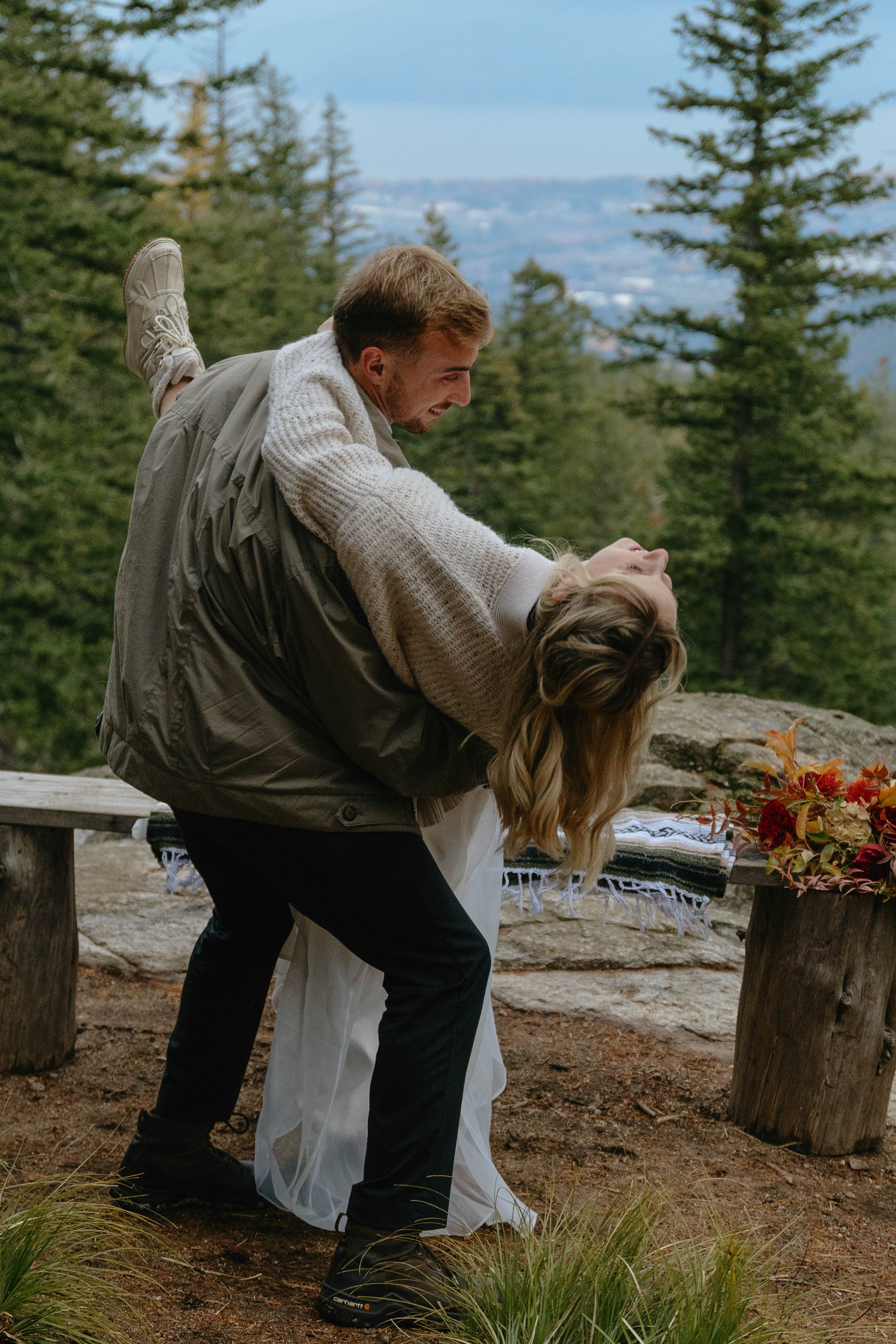 A couple is dancing outdoors in a forested area, with the man lifting the woman. The scene includes trees, a wooden bench, a blanket, and a decorated table with flowers.
