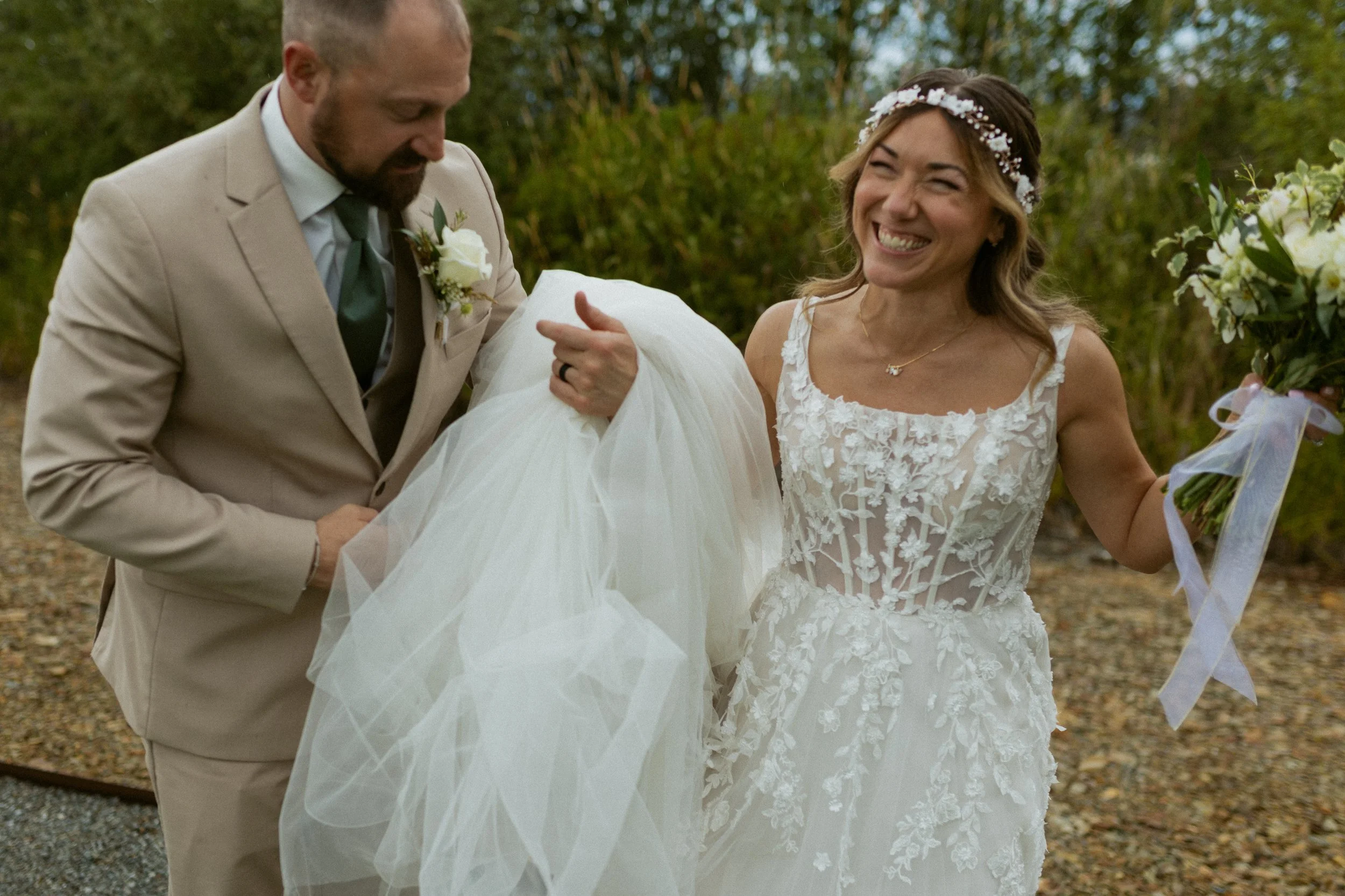 A bride with a floral headband and a lace wedding dress is smiling as a man in a beige suit helps carry her wedding dress. The bride holds a bouquet of white flowers.