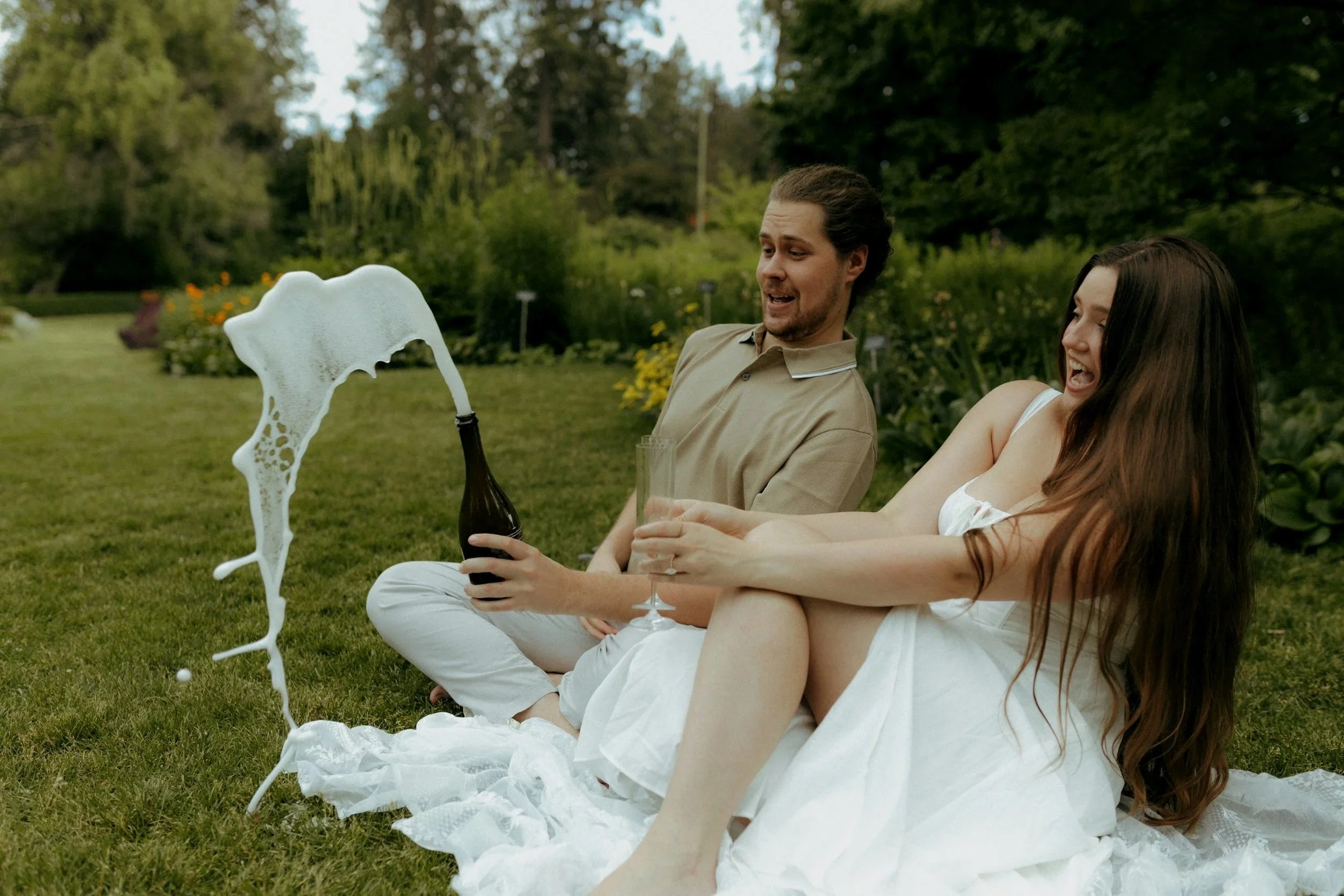 A man and woman sitting on the grass having a drink, with foam spilling out of the bottle, in a garden with trees and flowers.
