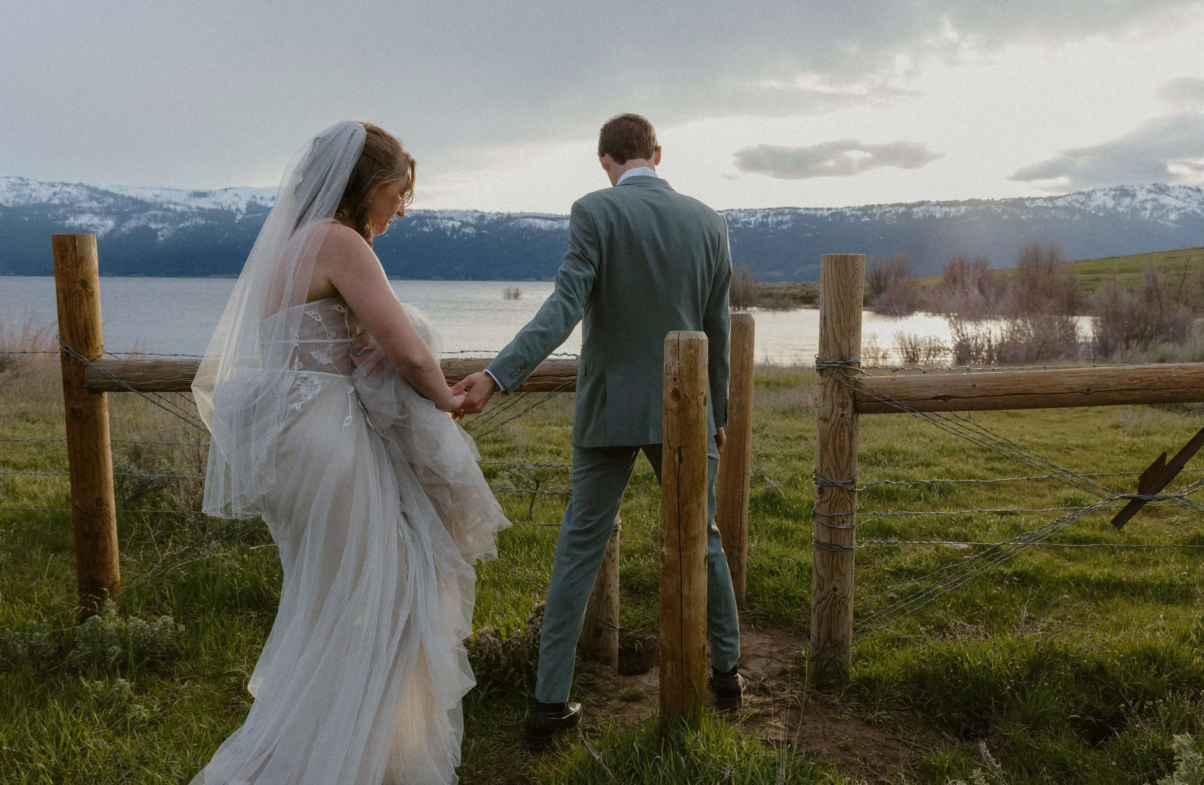 A bride and groom holding hands and walking by a wooden fence in a scenic outdoor landscape with a lake, mountains with snow, and cloudy sky in the background.