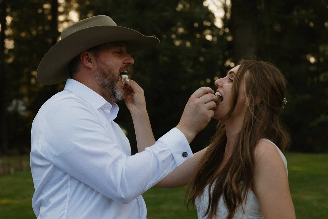 A man wearing a cowboy hat and white shirt feeds a woman in a white dress outdoors with trees in the background.