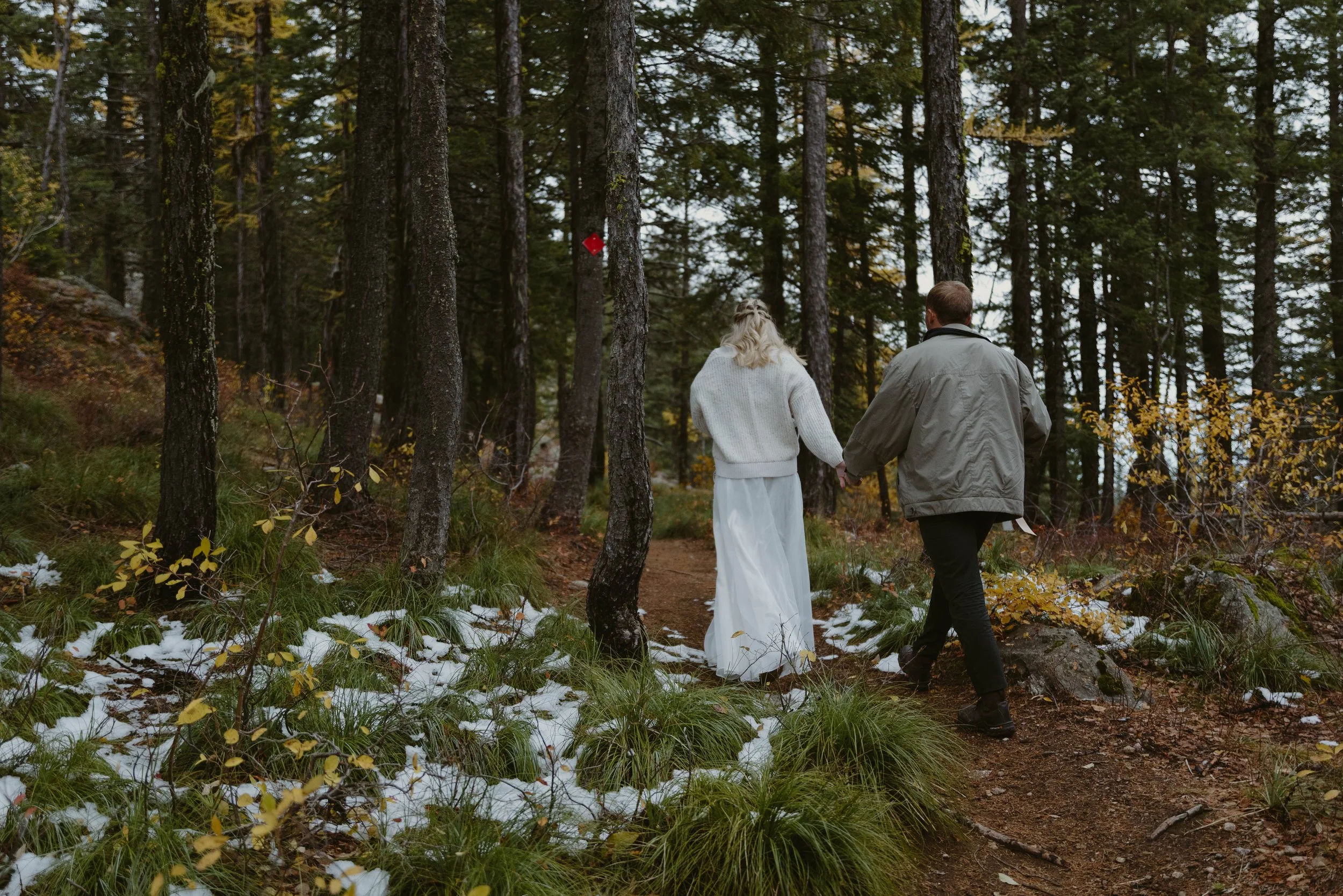 A couple holding hands walking in a forested area during autumn, with some snow on the ground.