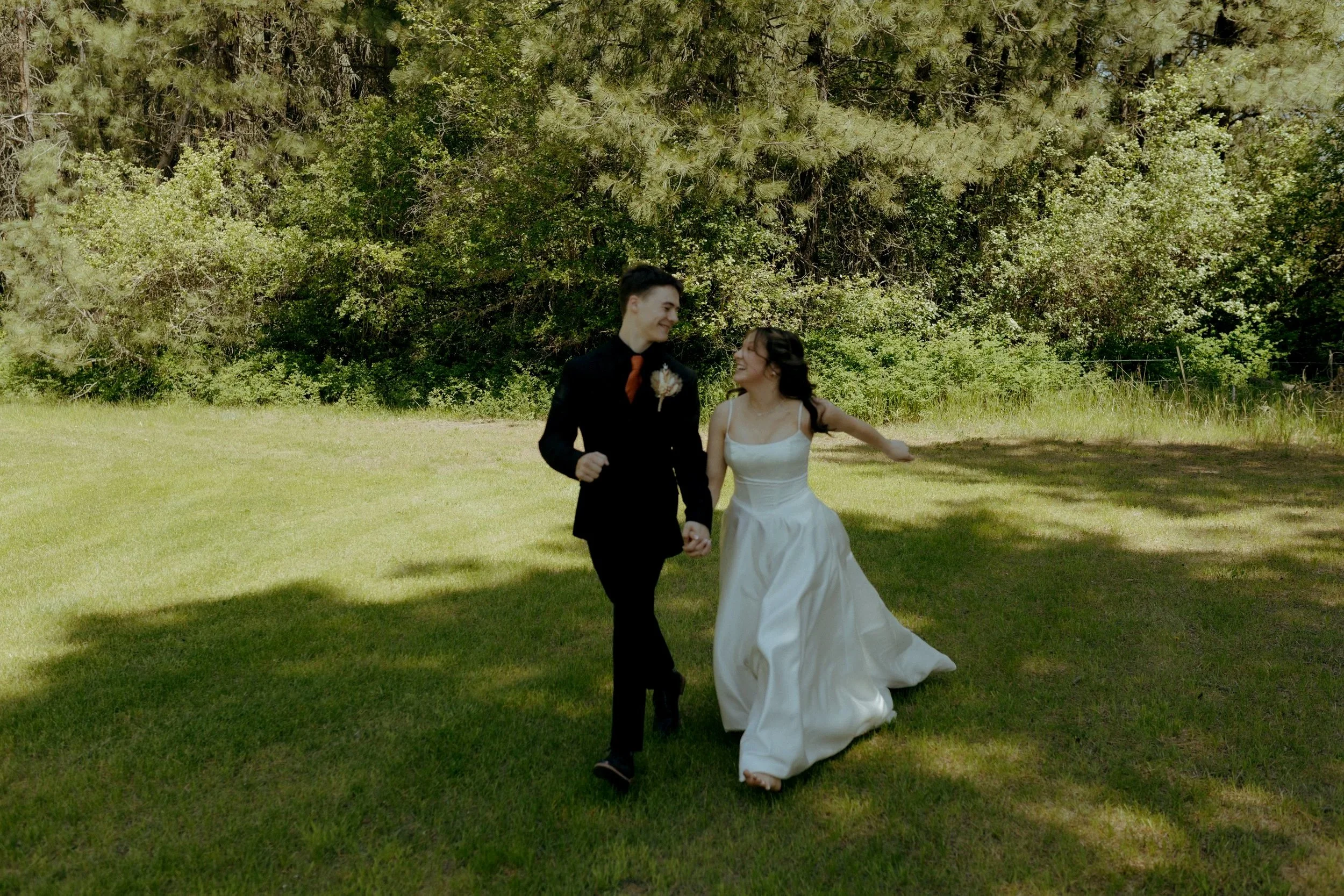 A smiling young man and woman holding hands and walking outdoors on a grassy field, with trees in the background on a sunny day.