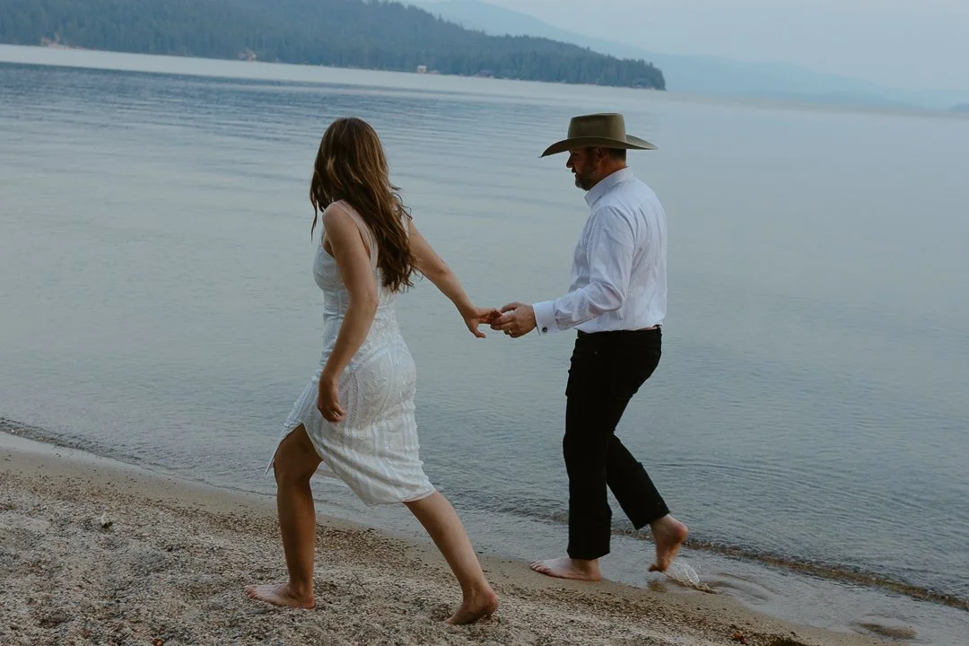 A man and woman holding hands and walking barefoot along the shoreline of a lake or ocean, with mountains in the background.