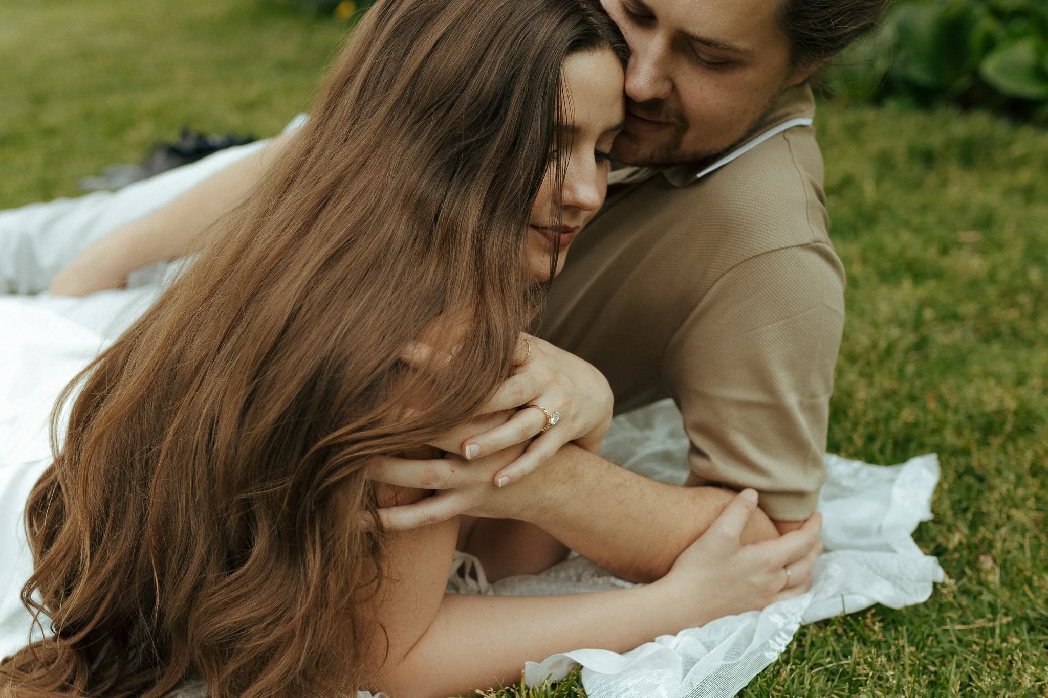 Romantic couple lying on a blanket in a grassy area, embracing closely with eyes closed, the woman has long brown hair and an engagement ring, the man has short facial hair and is wearing a tan shirt.