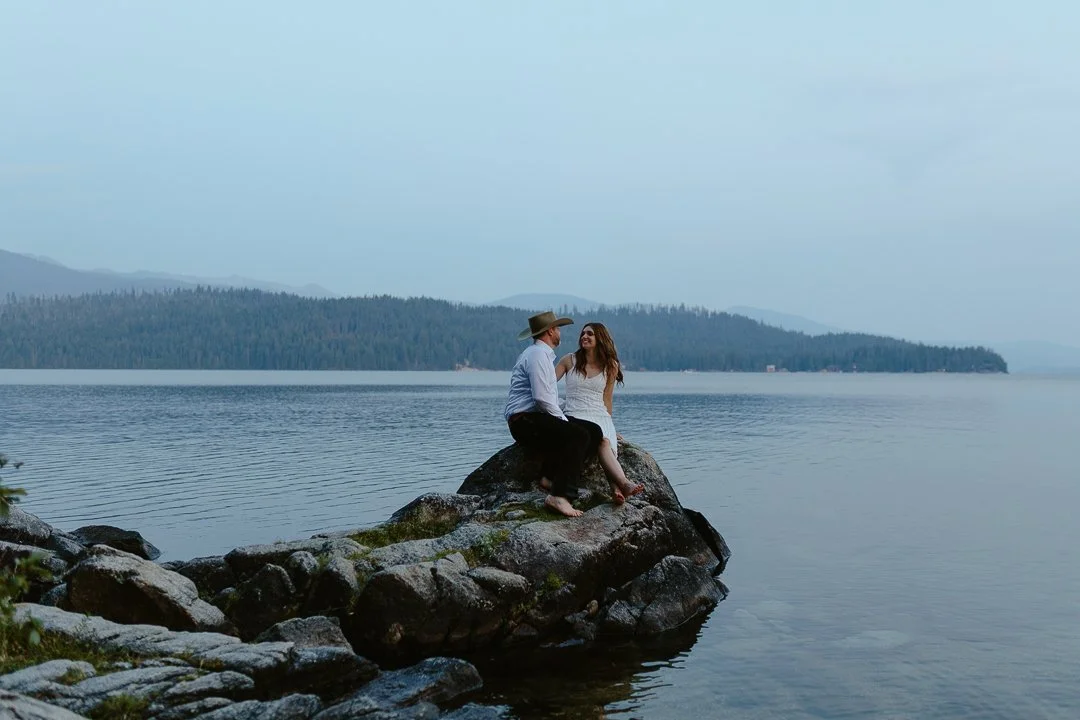 A couple sitting on a large rock at the edge of a lake, with forested hills in the background and a cloudy sky.