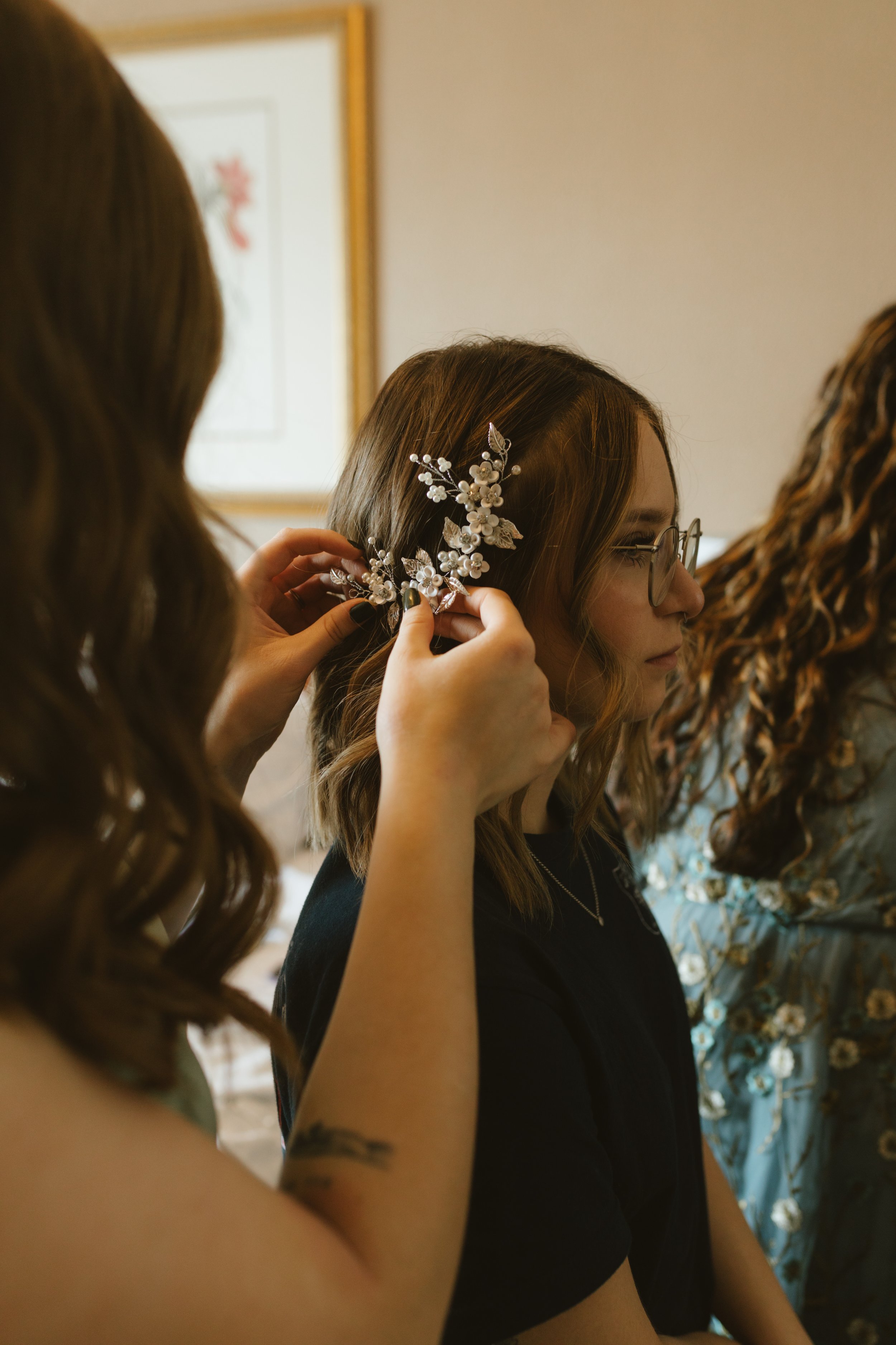 A woman with glasses and wavy hair getting a decorative floral hairpiece placed in her hair by another person.
